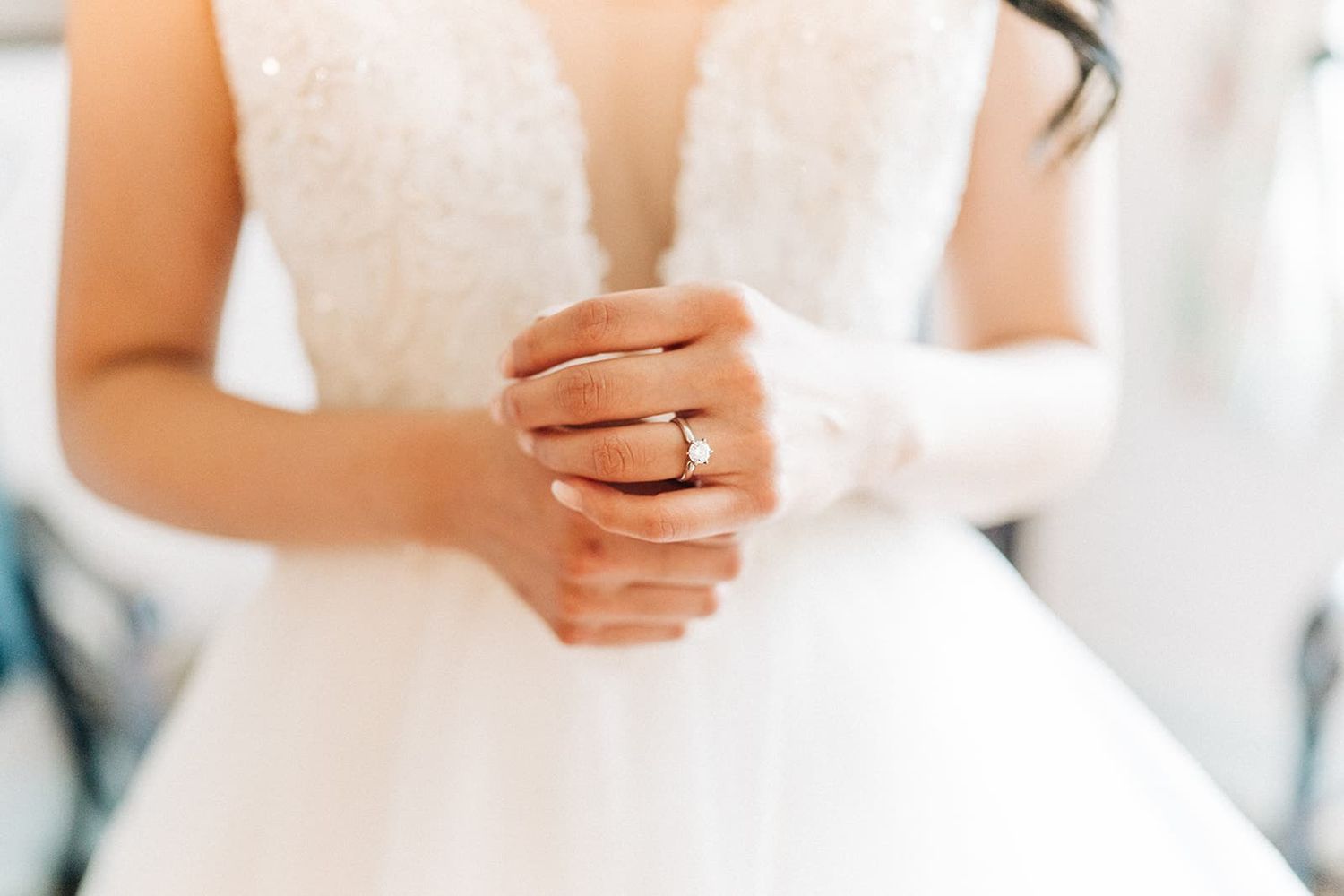 Bride in white lace wedding gown clasping hands showing gold diamond engagement ring.