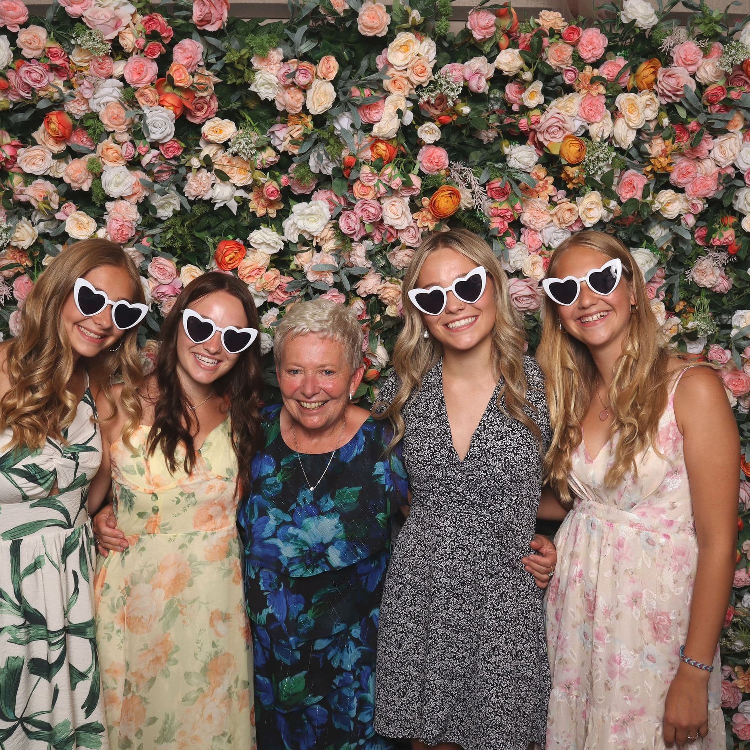 Five women pose in front of a floral wall backdrop, three wearing white heart-shaped sunglasses.