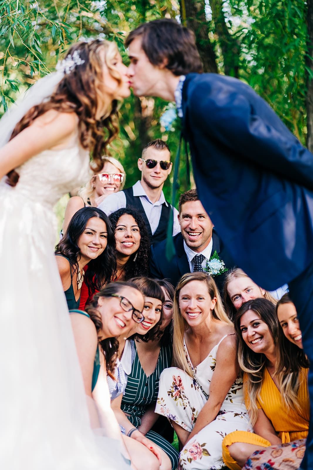 Wedding couple kissing while smiling guests pose for group photo outdoors under green trees.