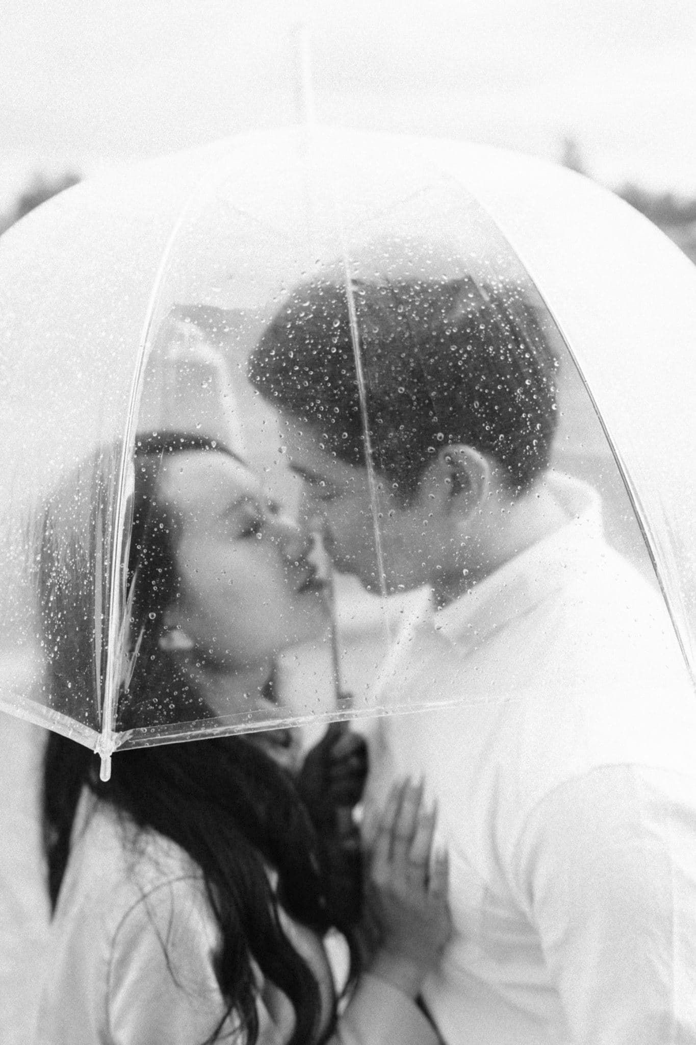Couple sharing a romantic kiss under a clear umbrella in the rain, black and white photo.