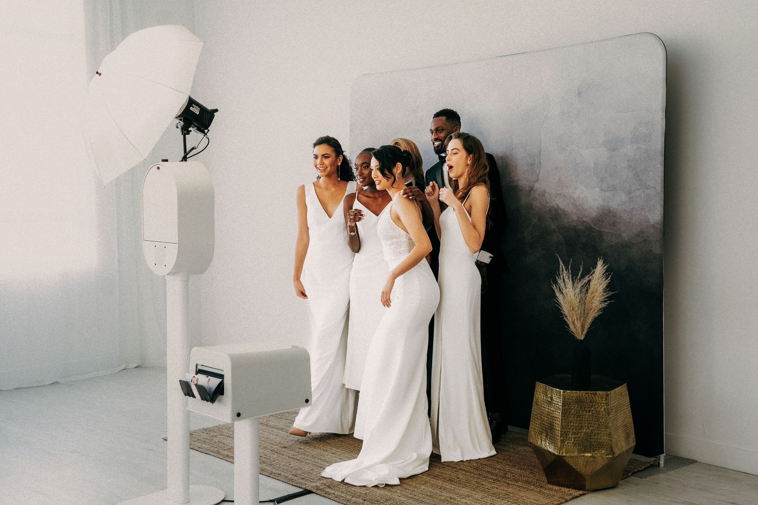 Four people wearing elegant white dresses pose together against a light gray wall in a studio setting.
