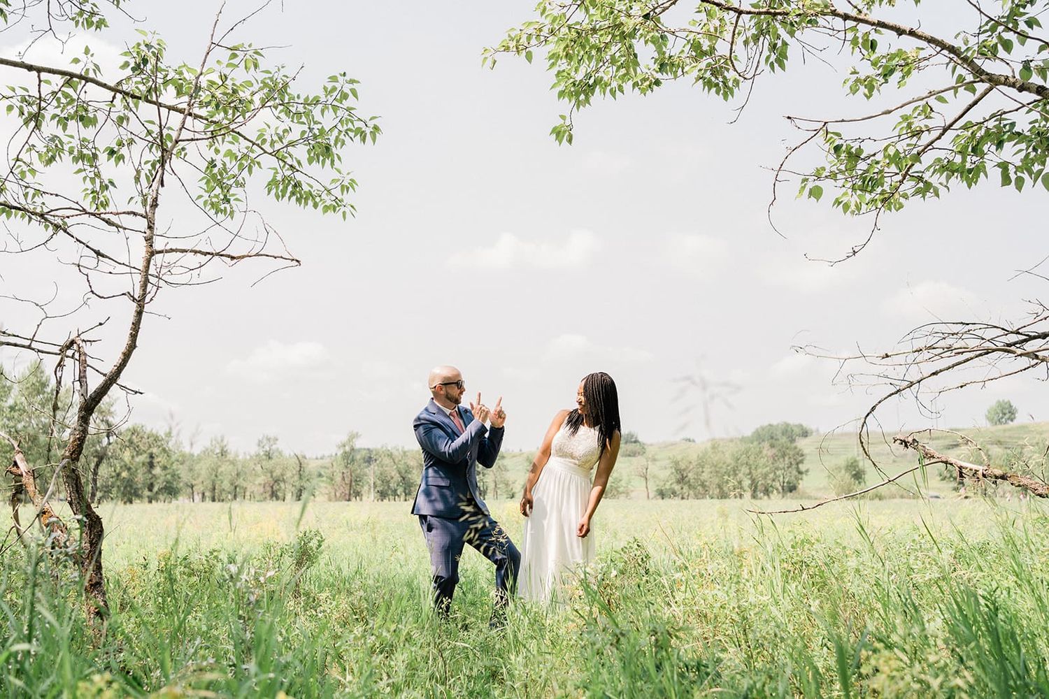 Couple dancing in a lush green meadow framed by trees on a bright summer day in a romantic outdoor setting.