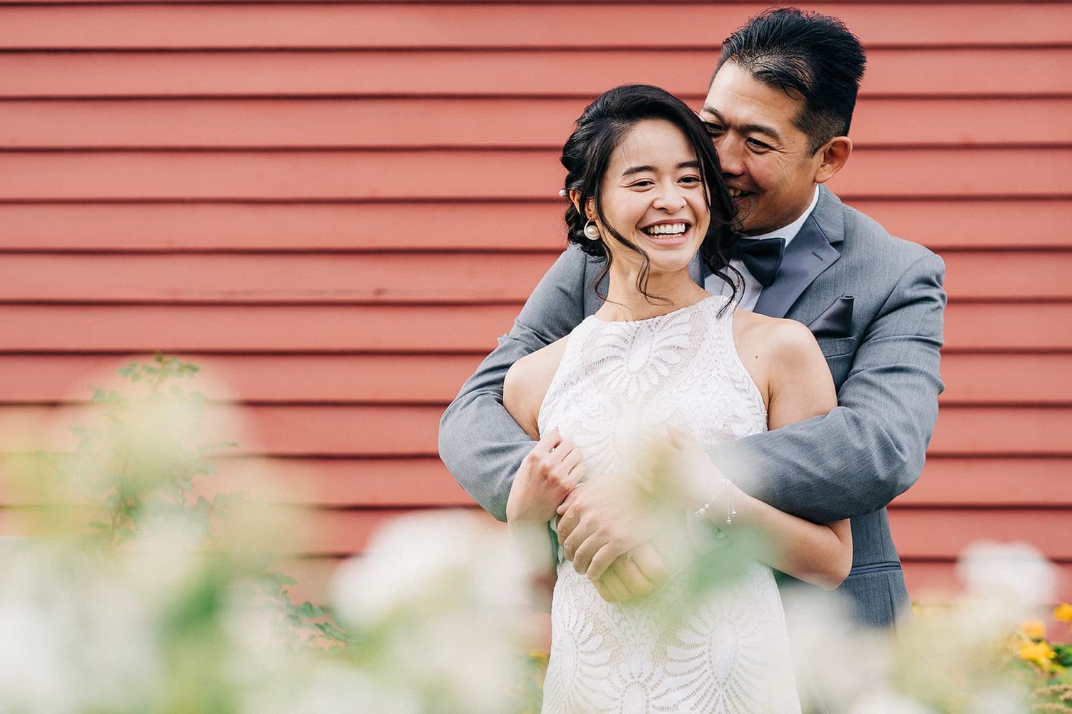 Happy Asian couple embracing in front of red barn wall, bride in white lace gown and groom in gray suit.