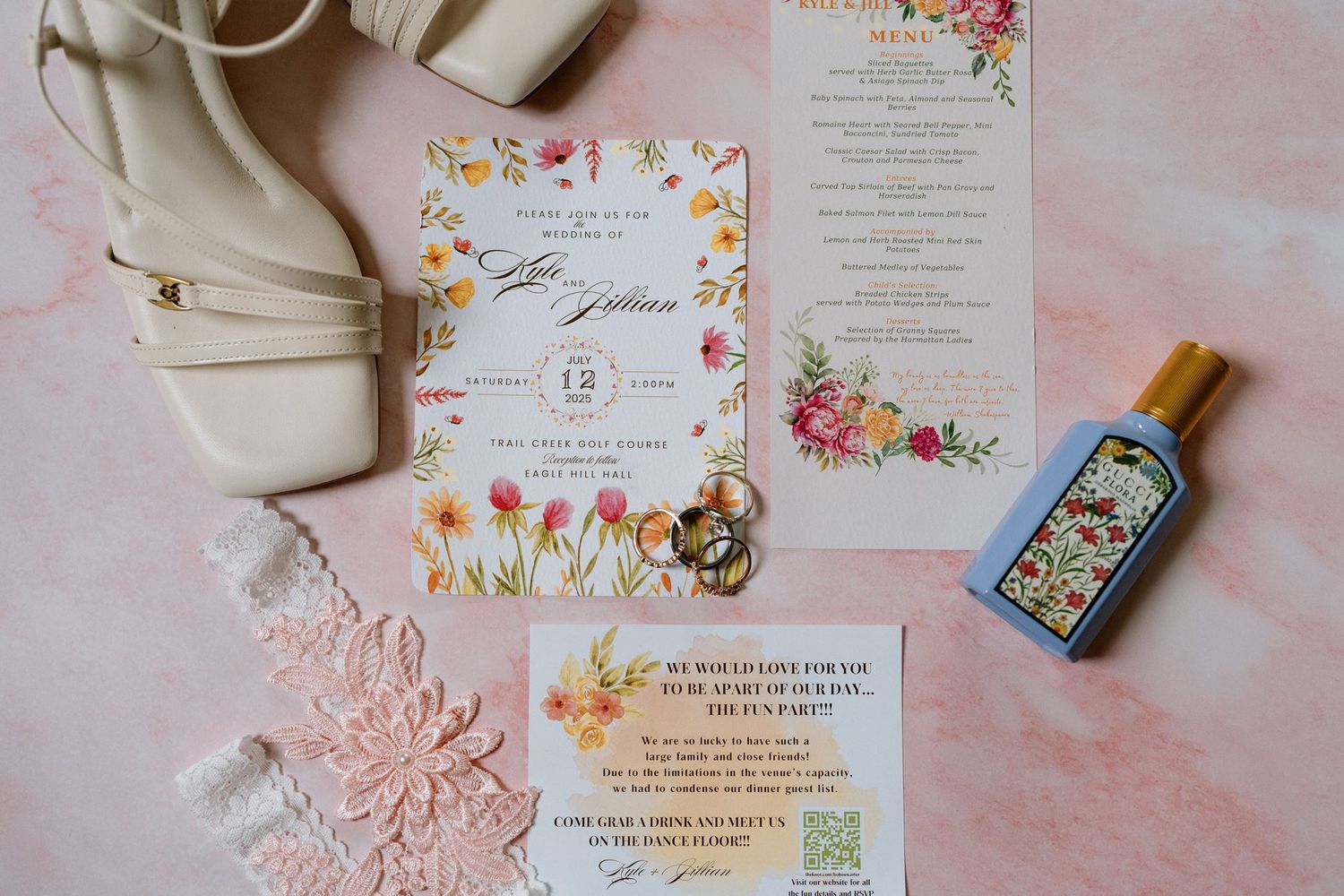 Floral wedding invitation suite with rings, white heeled sandals, lace details, and a Gucci perfume bottle on a pink background.