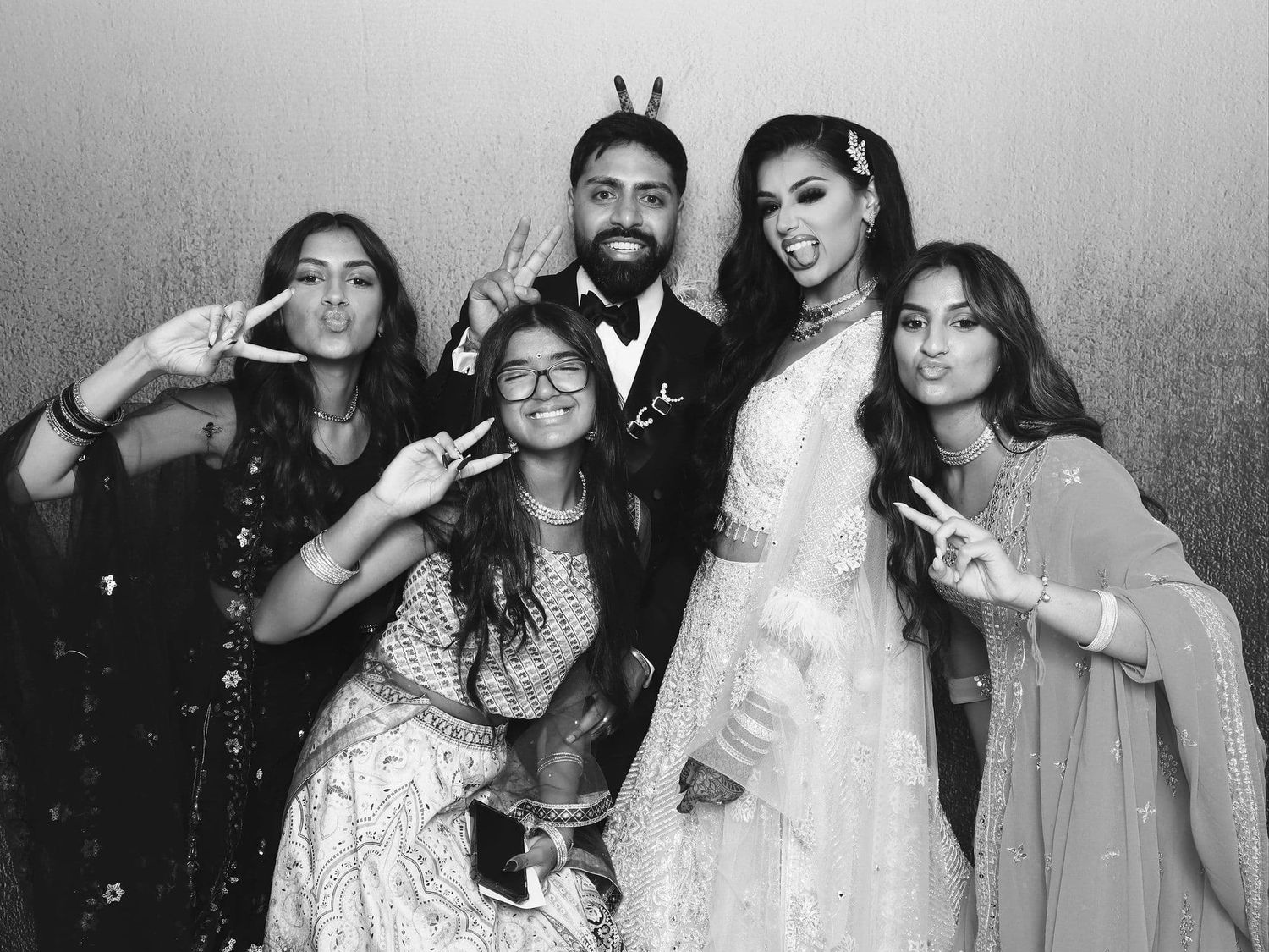 Group of five people in Indian formal wear pose with peace signs at a wedding celebration.