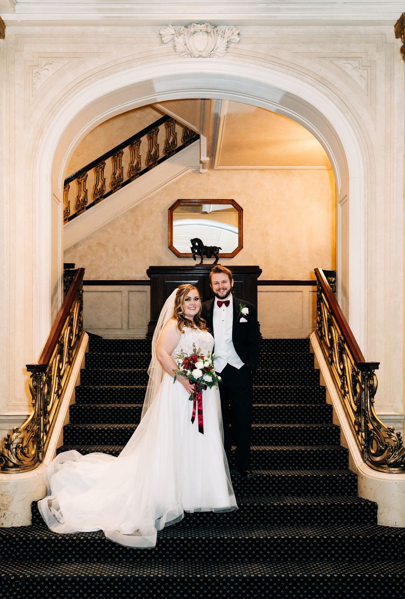 Bride and groom pose on elegant grand staircase with ornate gold railings, bride holding red and white bouquet.