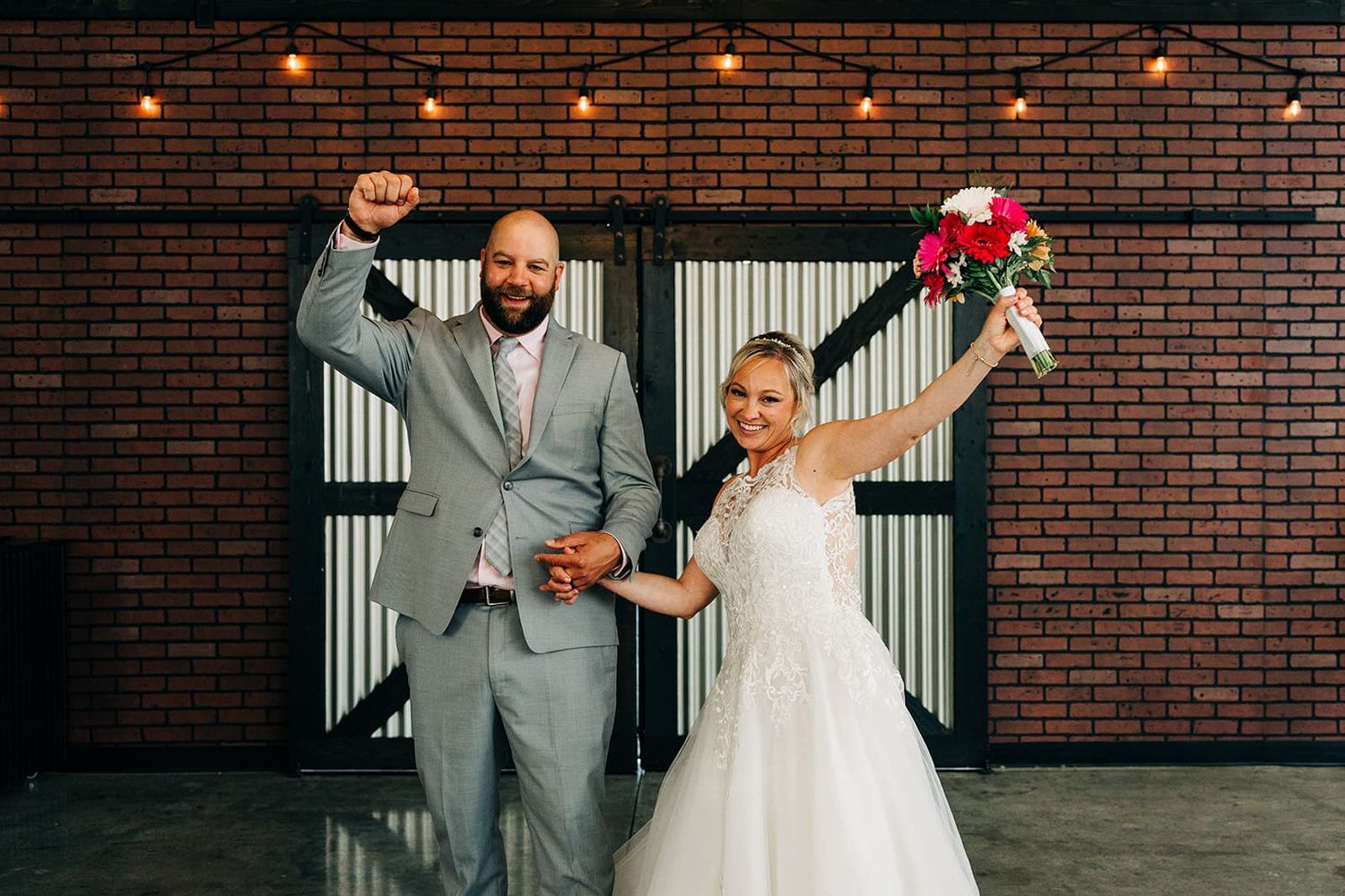 Excited newlyweds celebrate indoors, groom raising fist and bride lifting bouquet before a brick wall with string lights.