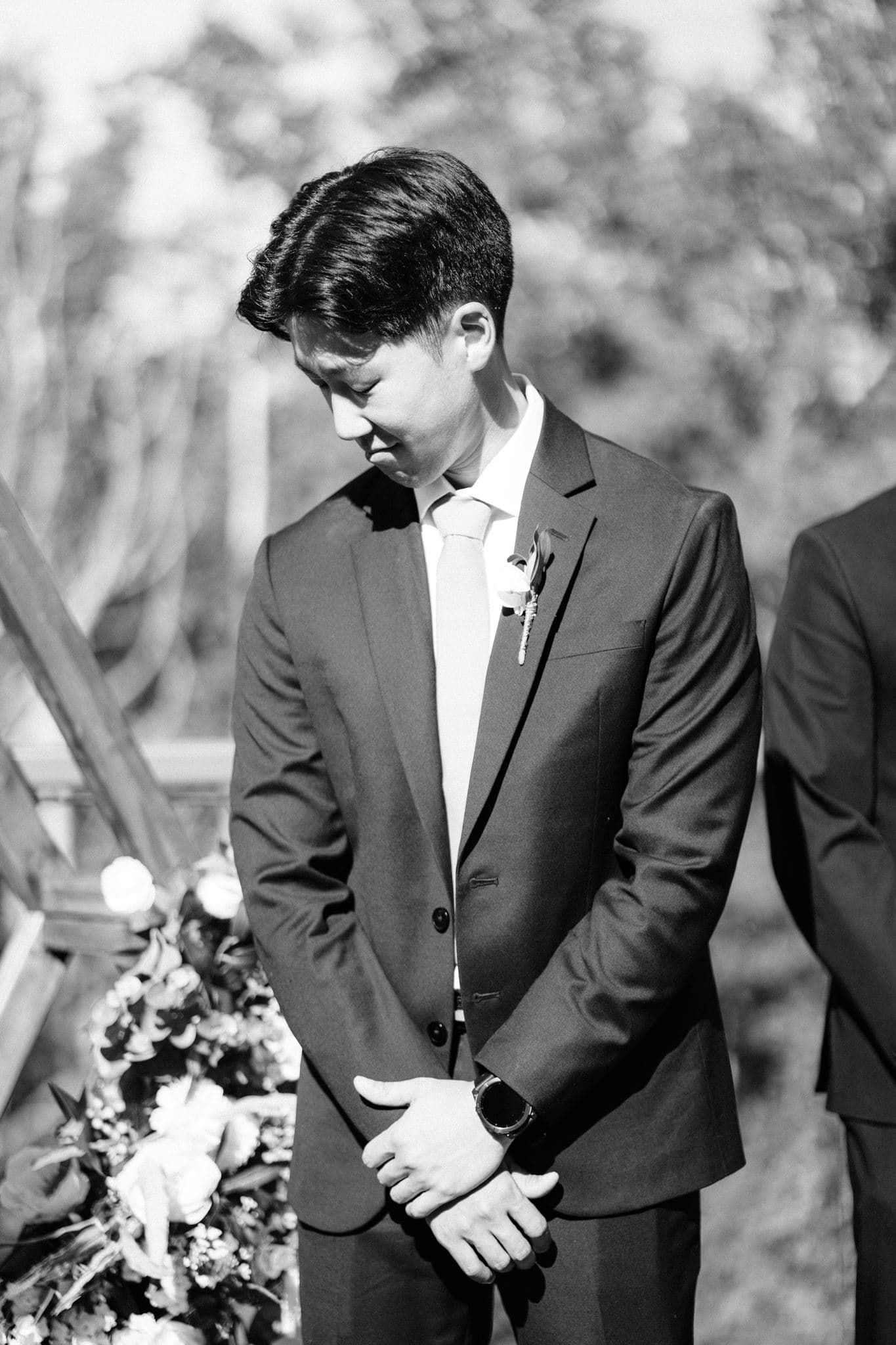 Black and white photo of a groom in a suit with boutonniere, hands clasped, looking down at a wedding ceremony.