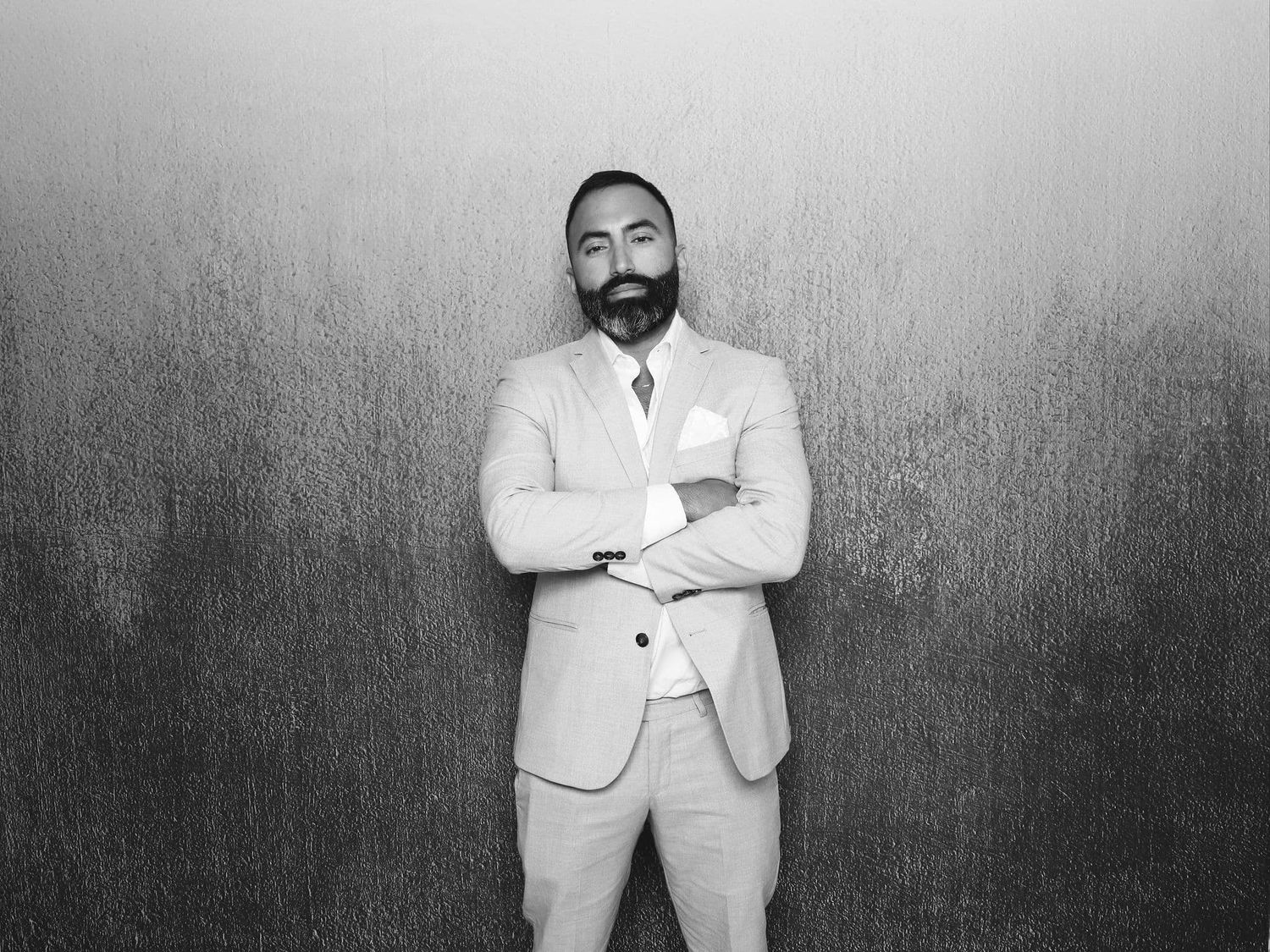 Man in light suit with crossed arms poses confidently against textured wall in black and white photo.