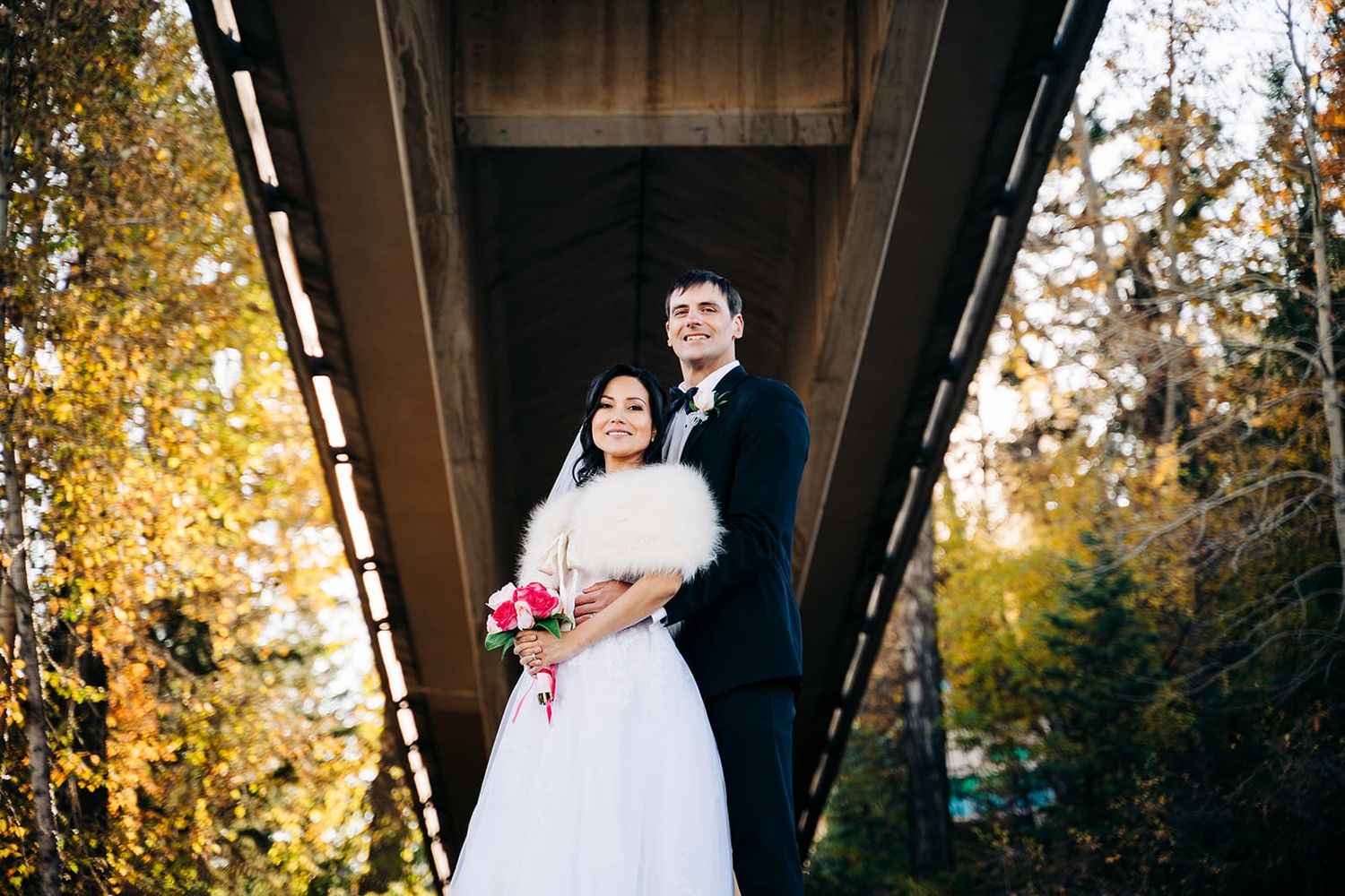 Bride and groom pose under a wooden bridge surrounded by autumn foliage, bride holding a pink bouquet.