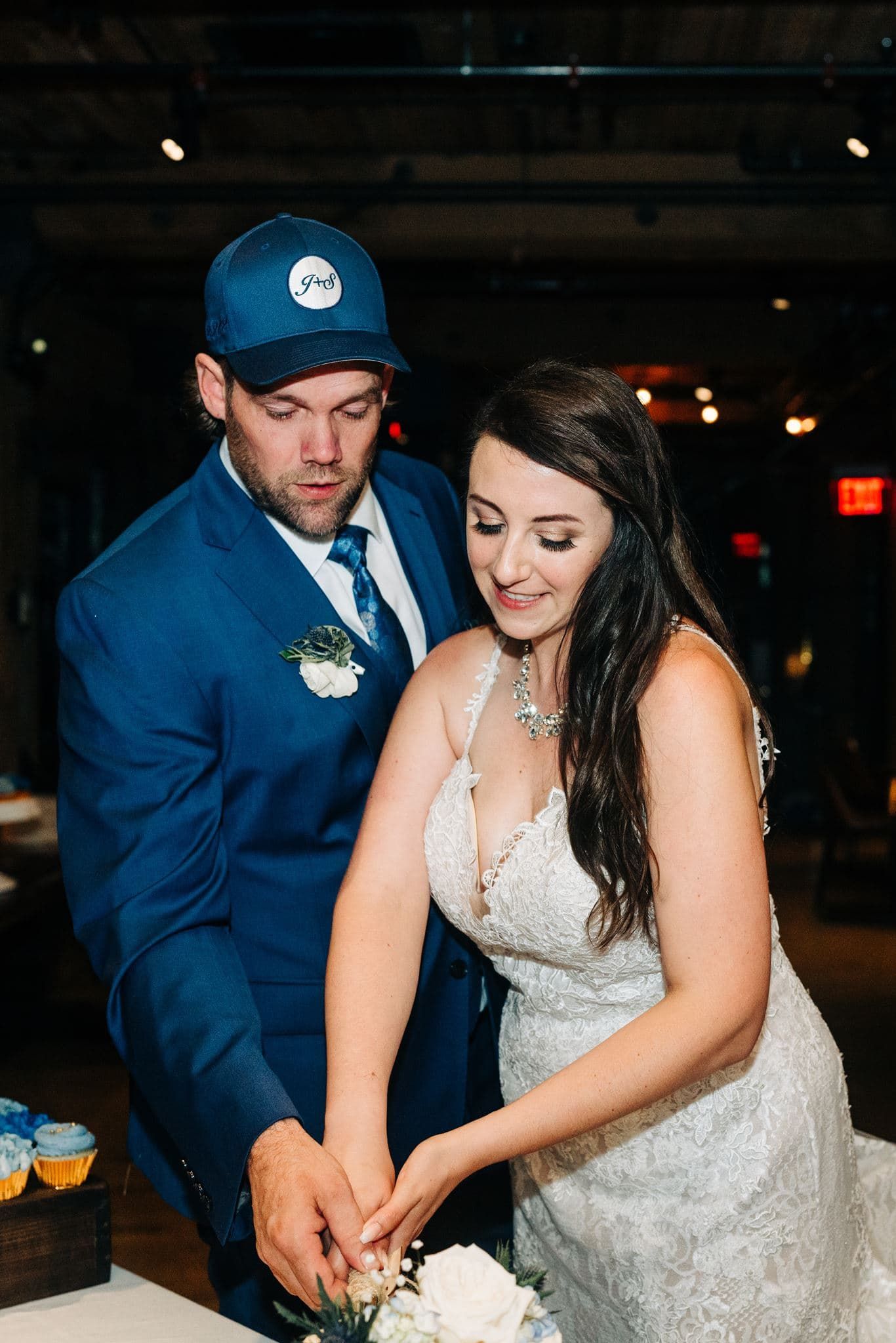 Bride and groom cutting wedding cake, groom in blue suit and trucker hat, bride in lace gown with floral bouquet.