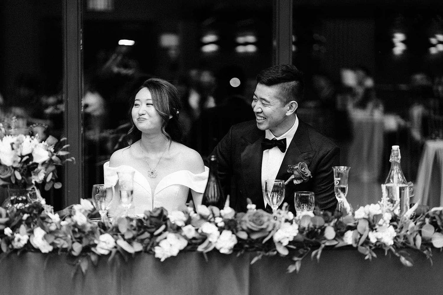 Black and white photo of a smiling Asian bride and groom in formal wedding attire seated at a floral-decorated reception table.