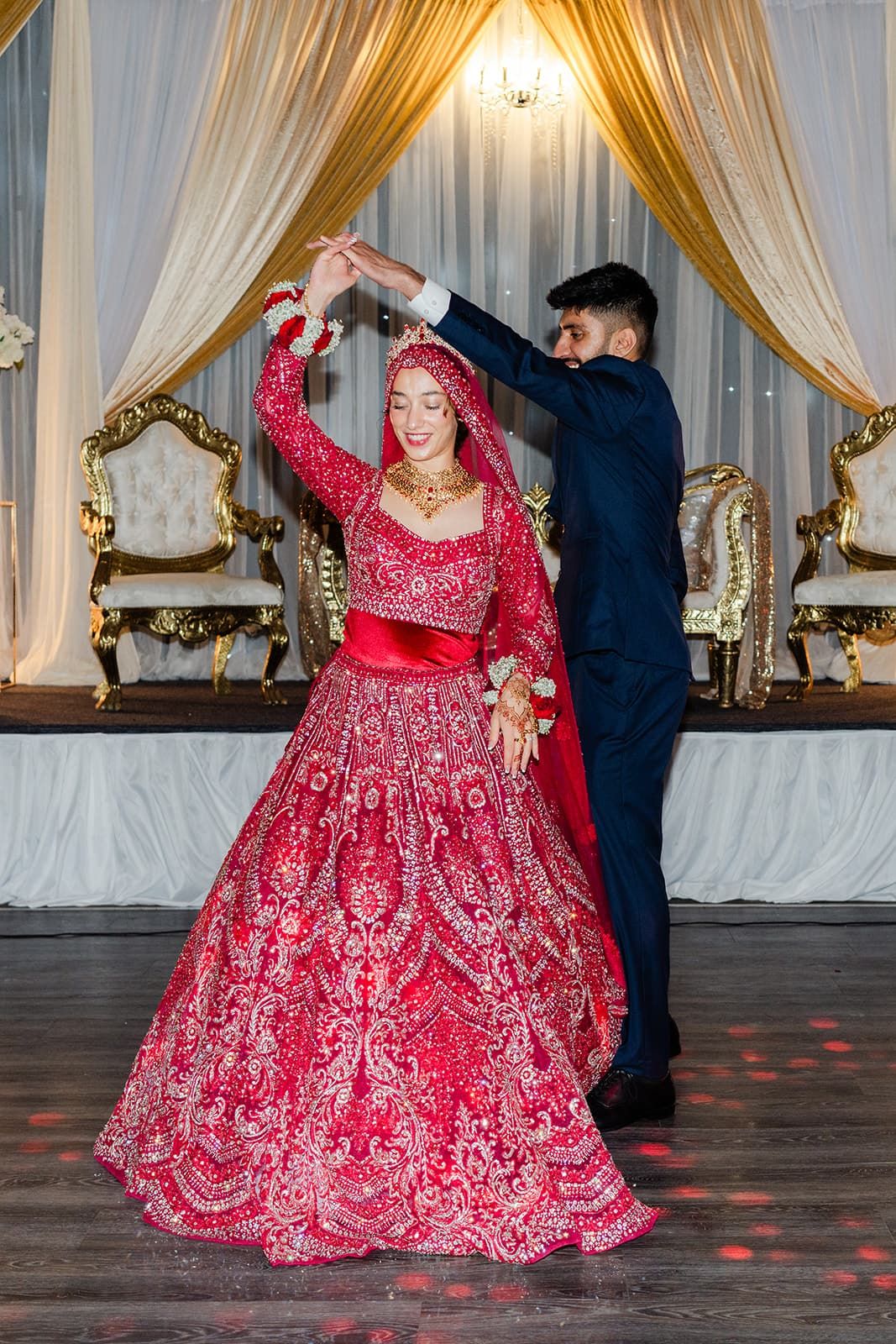 Bride in red lehenga spins during first dance with groom in navy suit at Indian wedding reception.