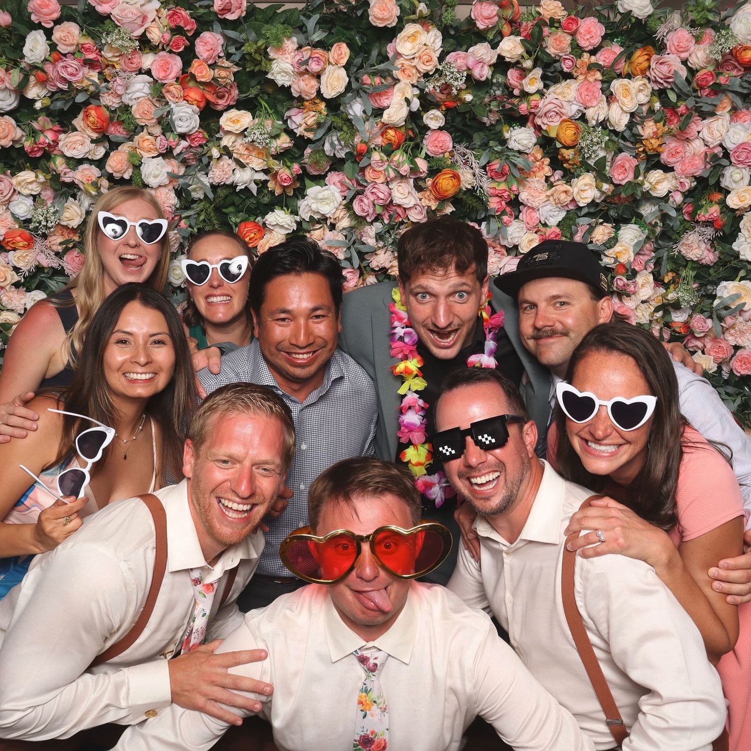 Group of ten joyful wedding guests pose with fun heart-shaped sunglasses in front of floral backdrop.