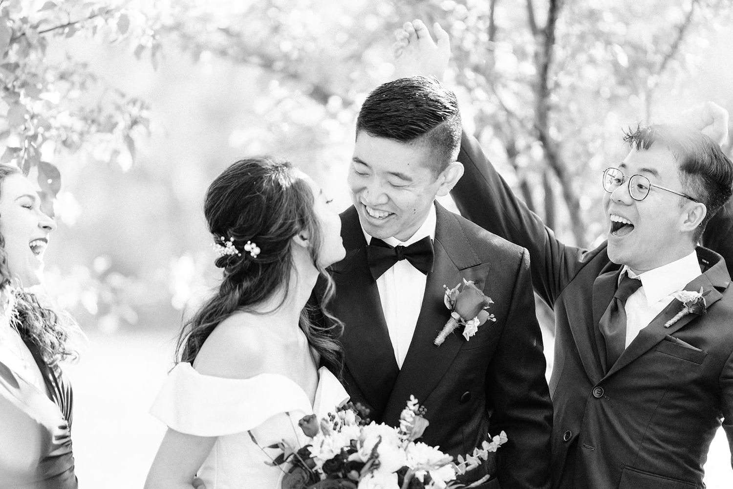 Black and white photo of a smiling groom in a tuxedo laughing with wedding guests outdoors.