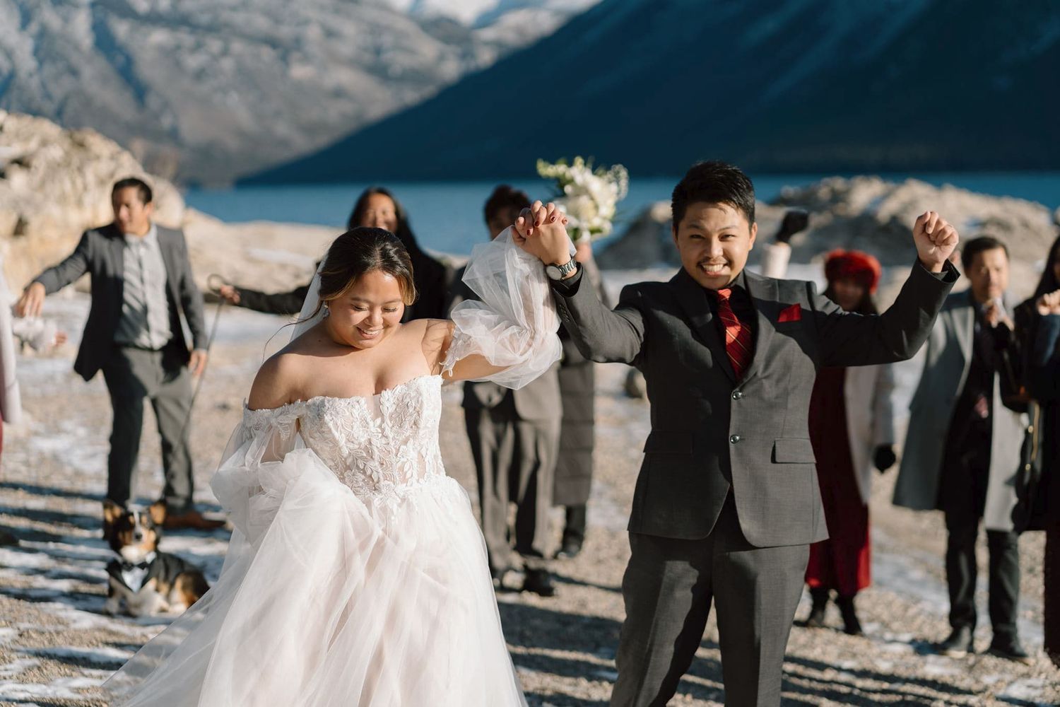 Bride and groom celebrate joyfully with wedding party on snowy mountain lakeside in winter elopement ceremony.