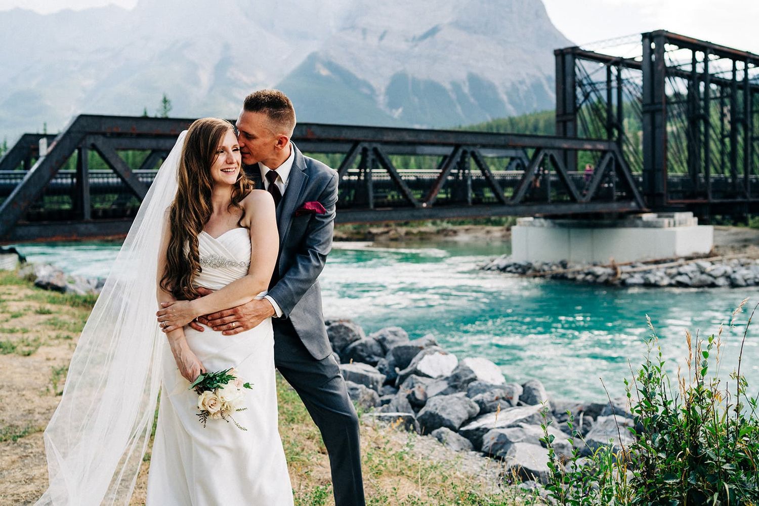 Bride and groom embrace by a turquoise river with a steel bridge and mountain backdrop on their wedding day.