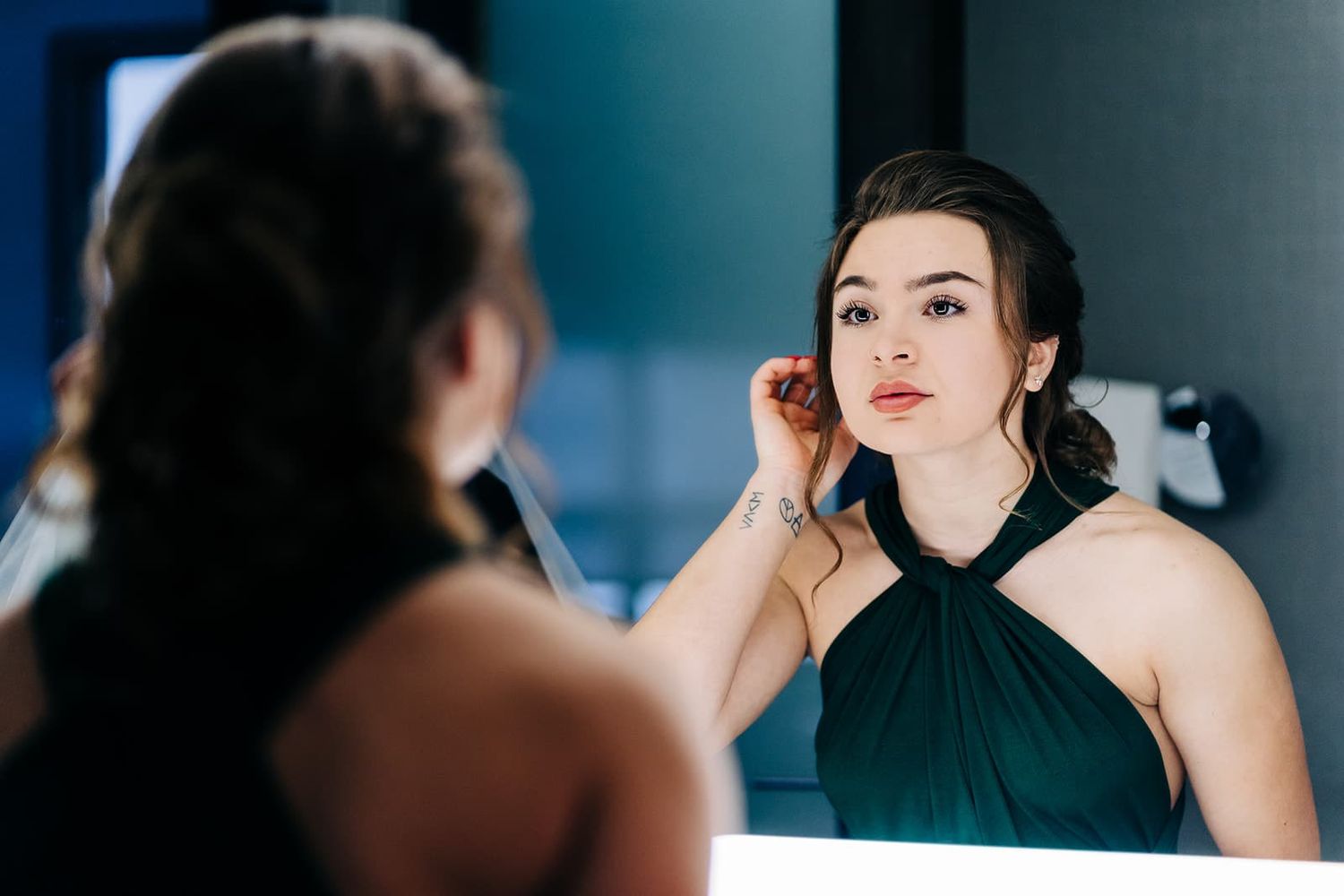 Young woman in elegant green halter dress adjusting earring while looking in illuminated bathroom mirror.