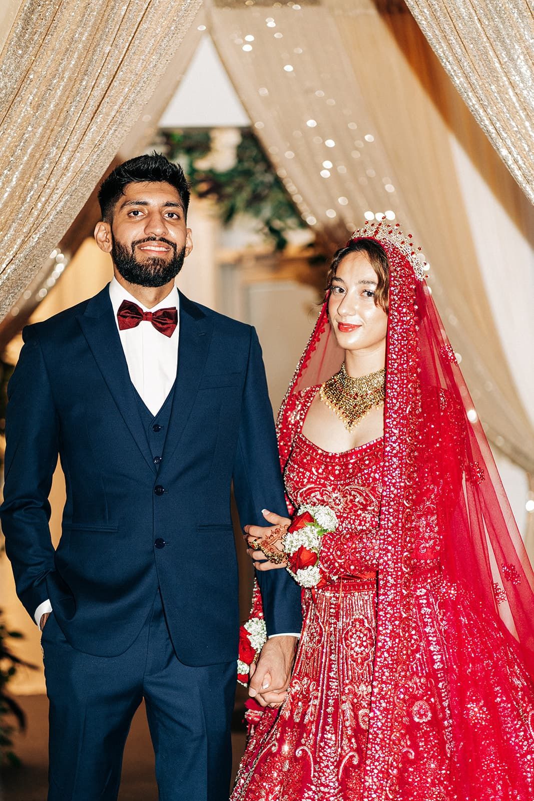 Indian bride and groom hold hands in traditional red lehenga and navy suit under golden mandap.