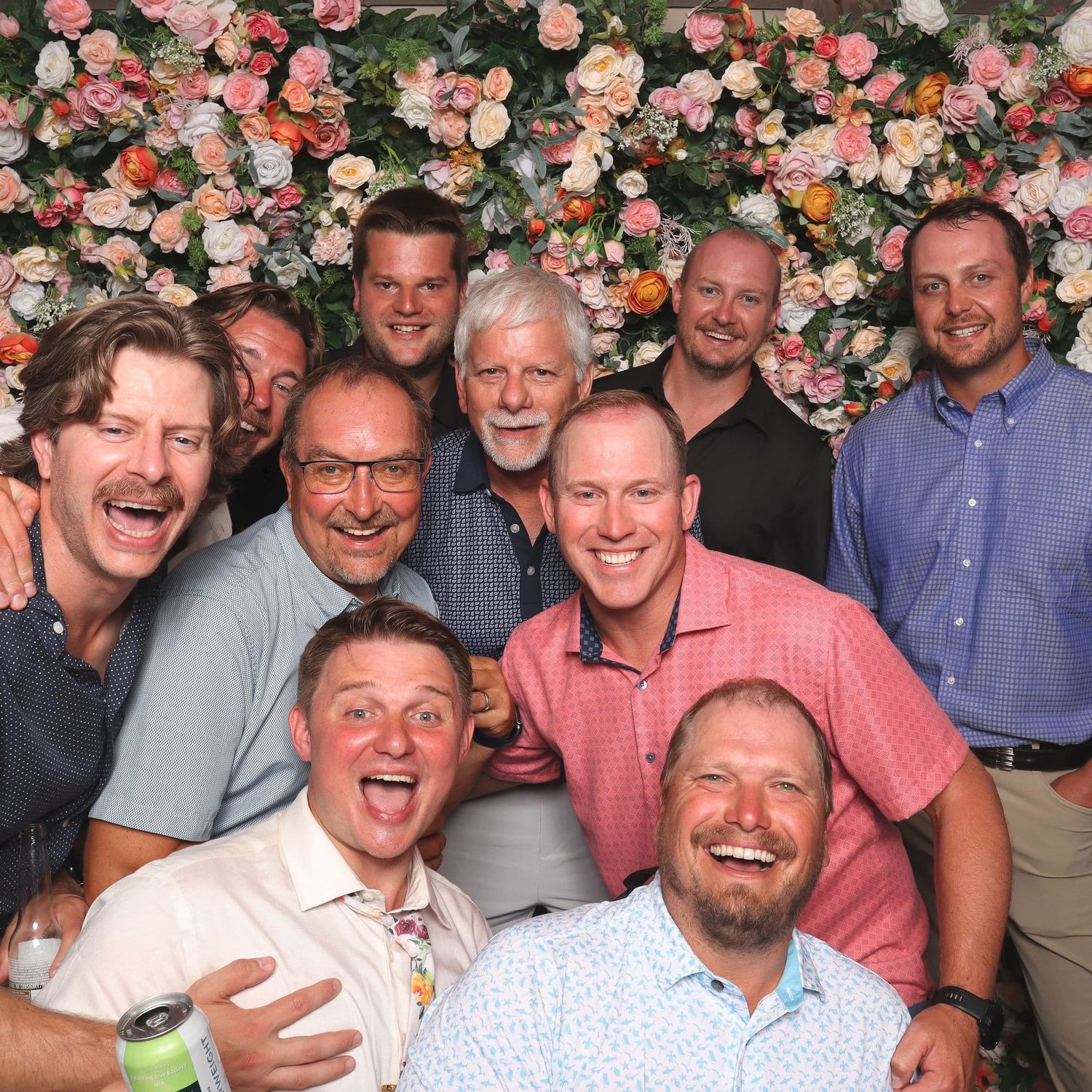 A large group of smiling men pose together in front of a floral wall backdrop.