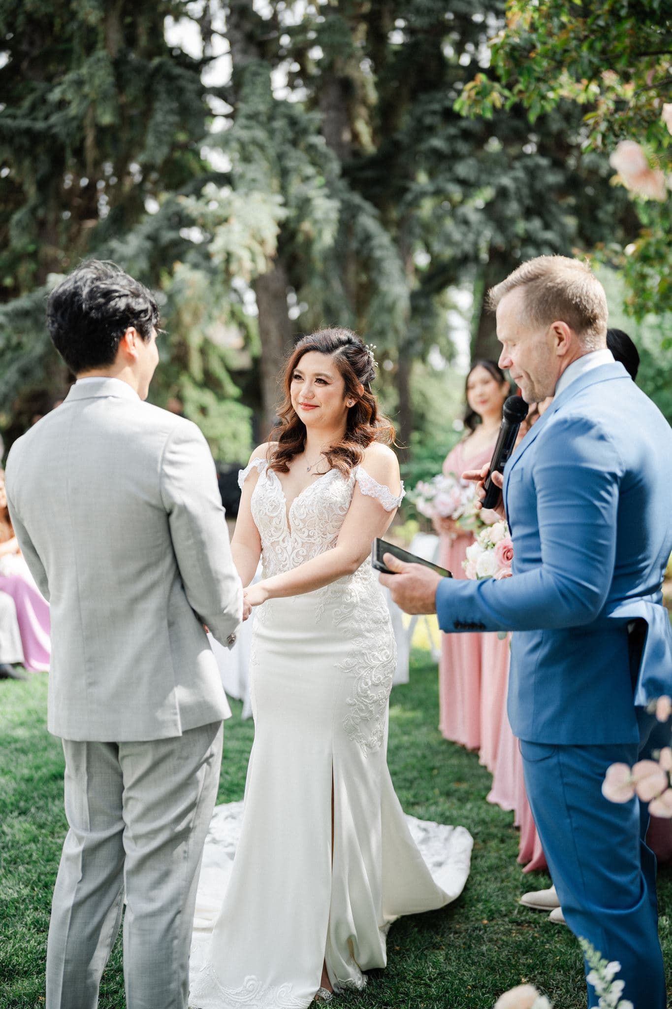 Bride and groom exchange vows outdoors with officiant in blue suit, surrounded by lush trees and bridesmaids.