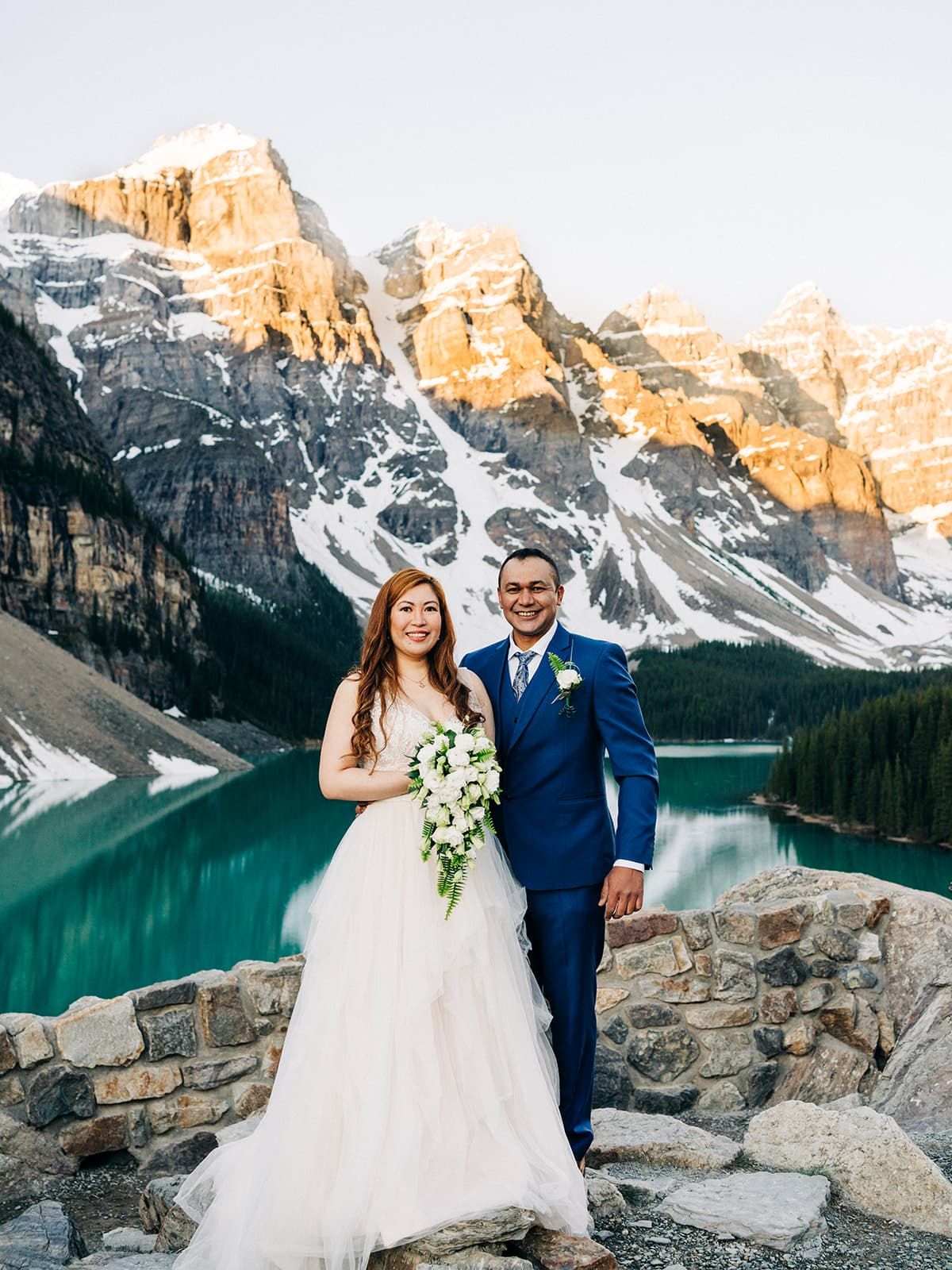 Bride and groom pose at Moraine Lake, Banff, with turquoise water and snow-capped Rocky Mountains at golden hour.