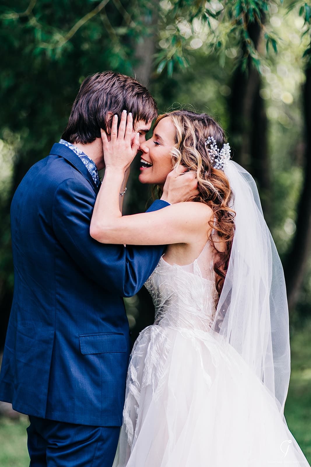 A joyful bride in a white gown and veil laughs as she holds the groom's face during an outdoor wedding ceremony.