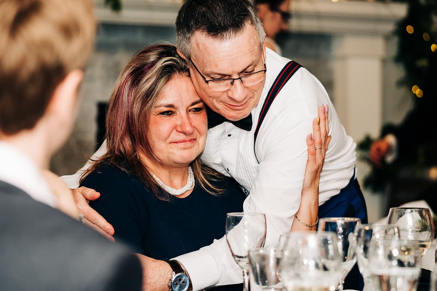 Emotional mother crying while being comforted by a man in suspenders at a wedding reception.