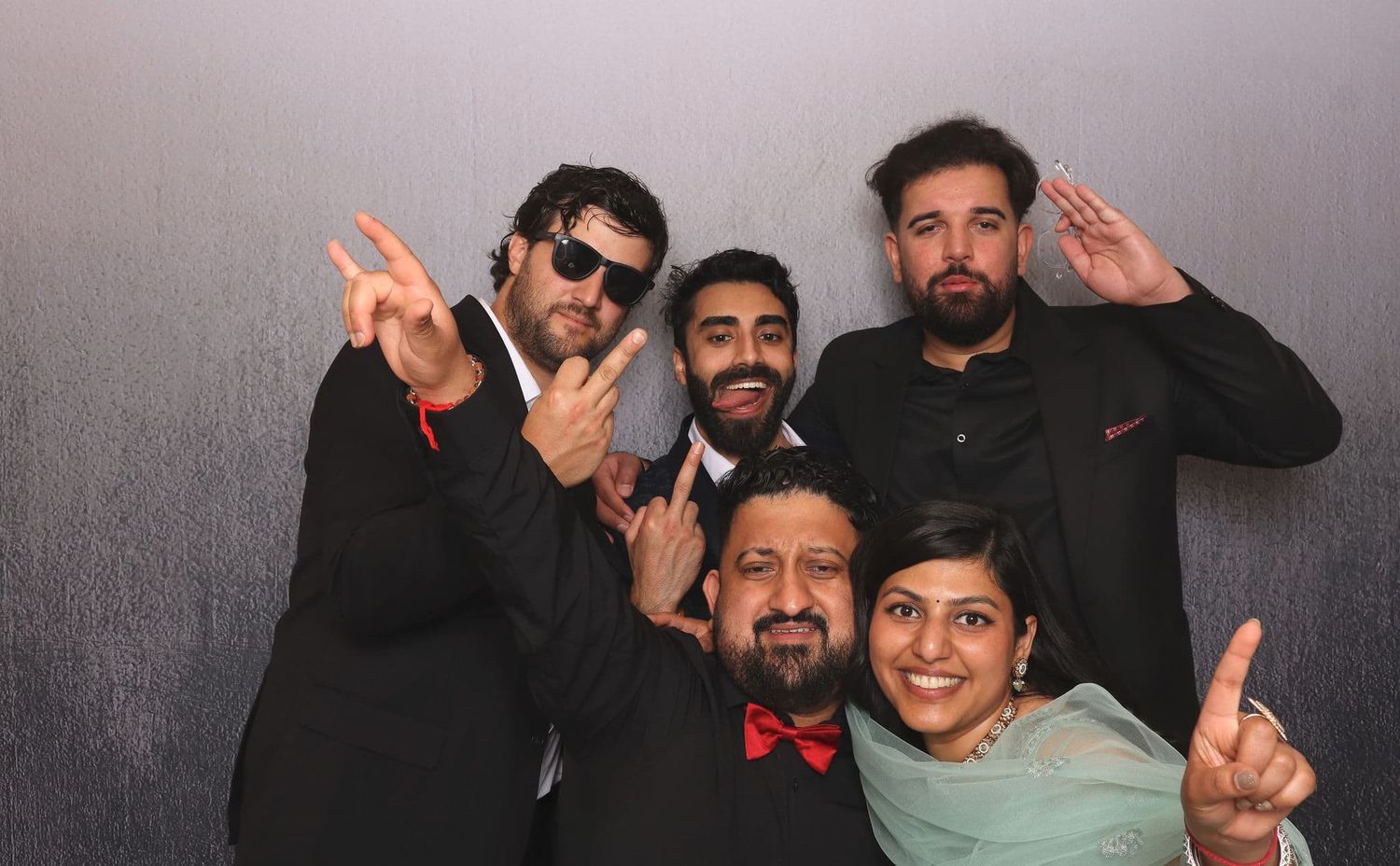 Five men in formal black suits making fun poses in a wedding photo booth.