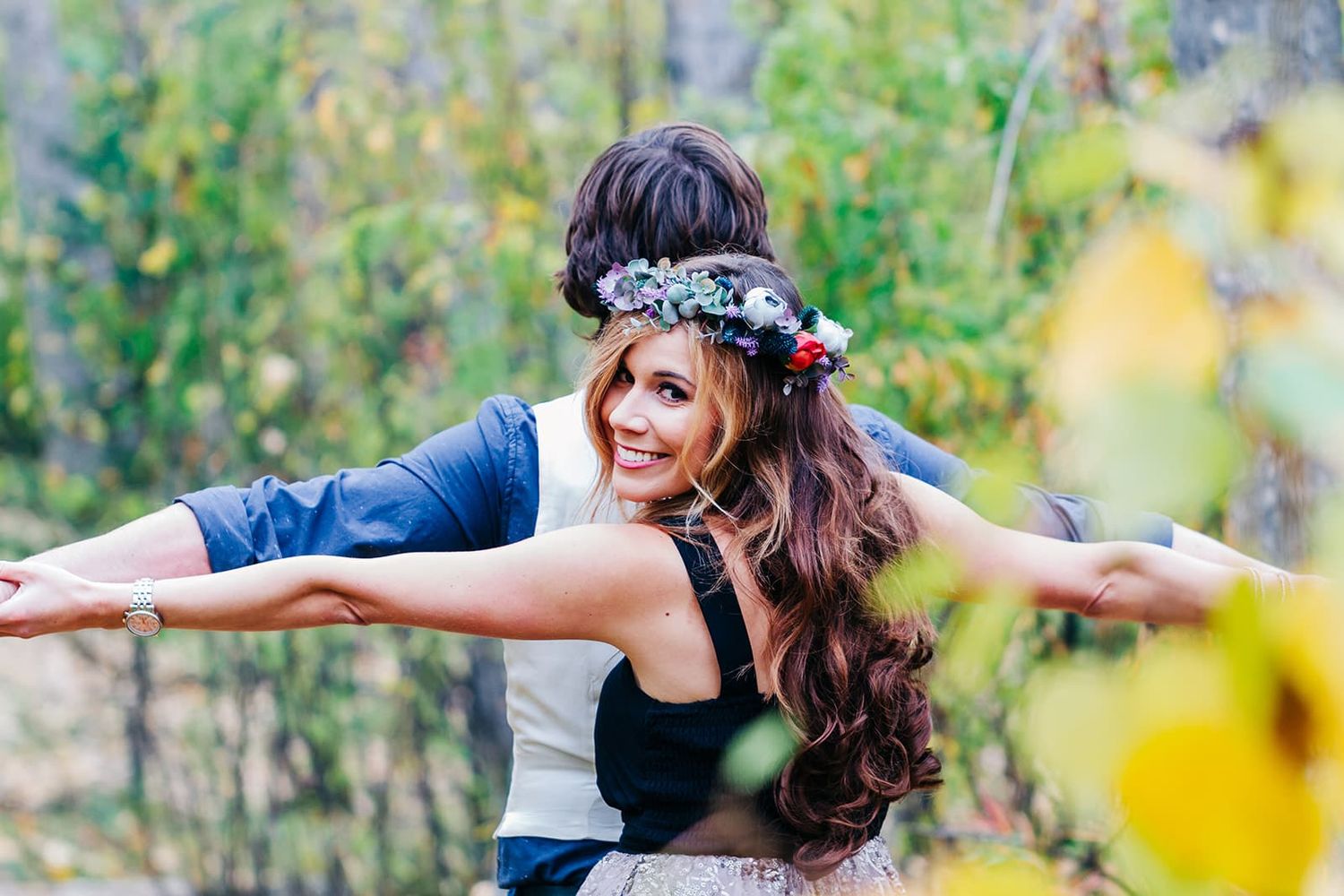 Woman with floral crown smiling outdoors, arms outstretched, with child on her back among autumn foliage.