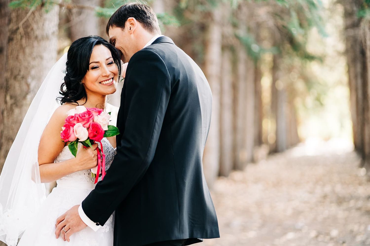 Bride and groom embracing outdoors among trees, bride holding pink bouquet and smiling in white gown and veil.