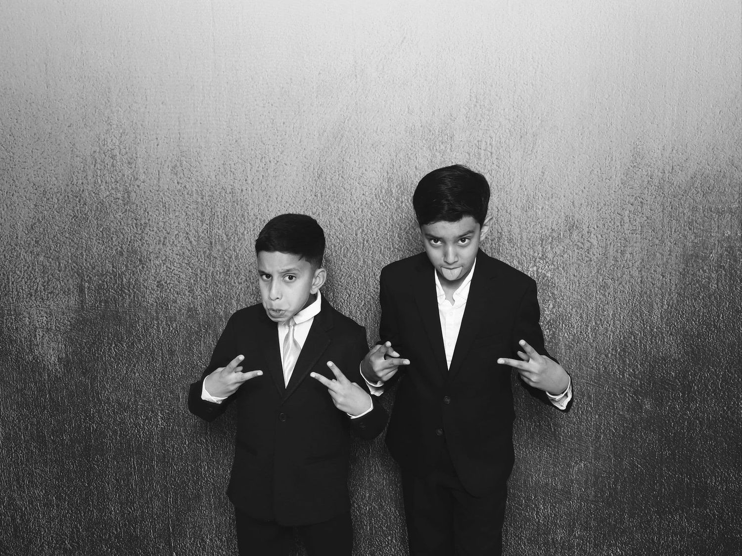 Two young boys in black suits make peace signs in a black and white portrait photo.