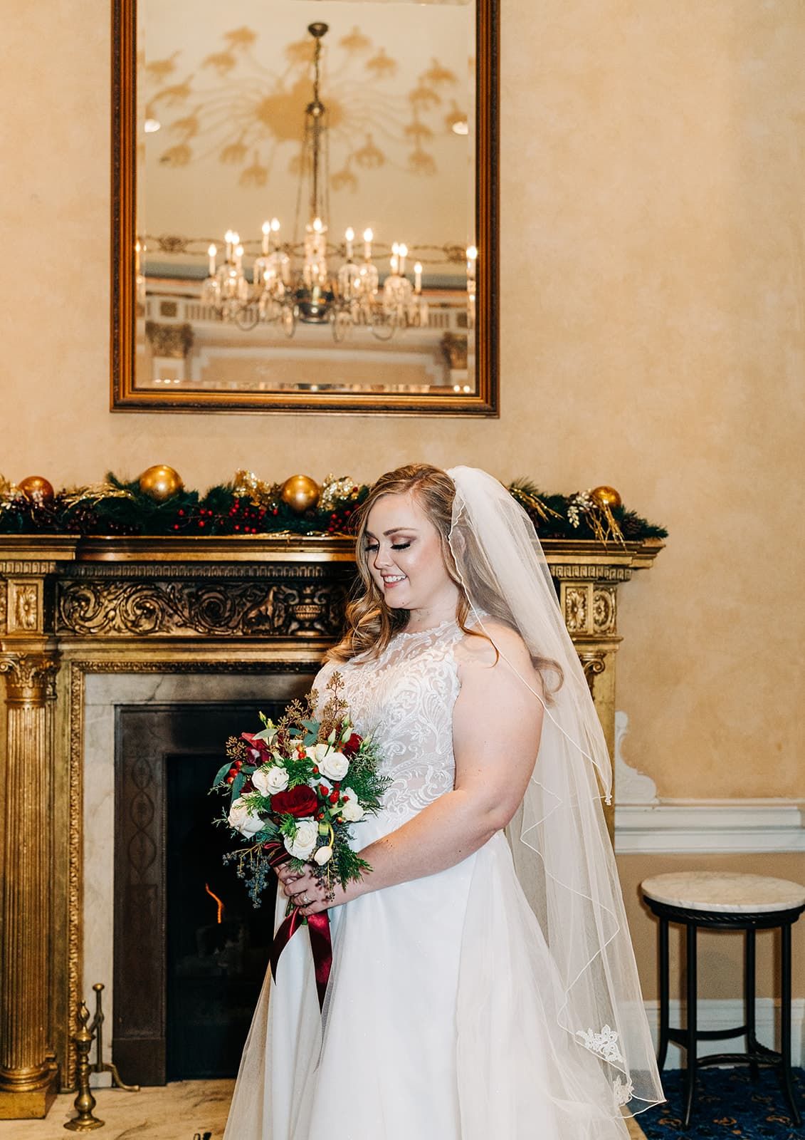 Bride in white gown holding red and white bouquet standing by ornate fireplace with chandelier.