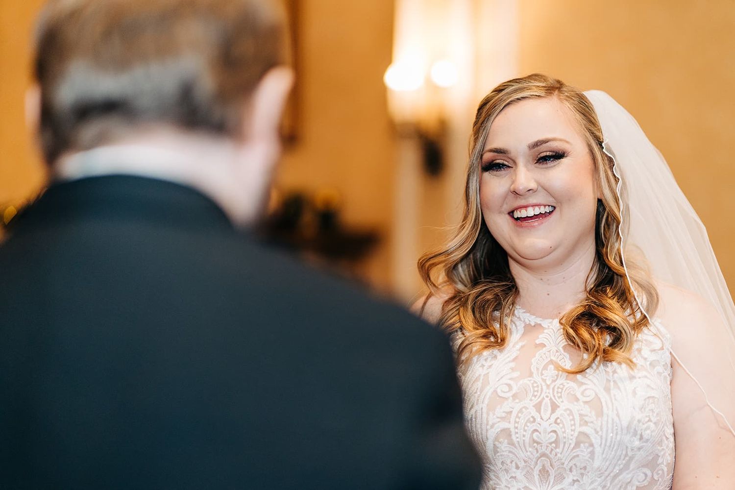 Smiling bride in lace wedding dress and veil exchanging vows with groom during indoor wedding ceremony.