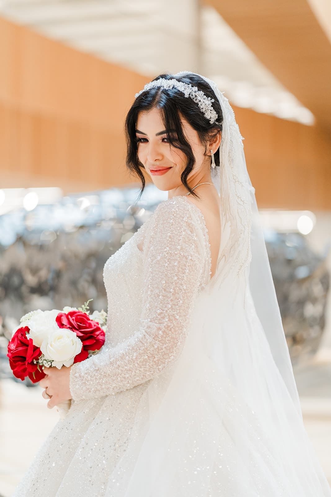 Bride in beaded white gown and veil holds red and white rose bouquet, wearing crystal hair piece.