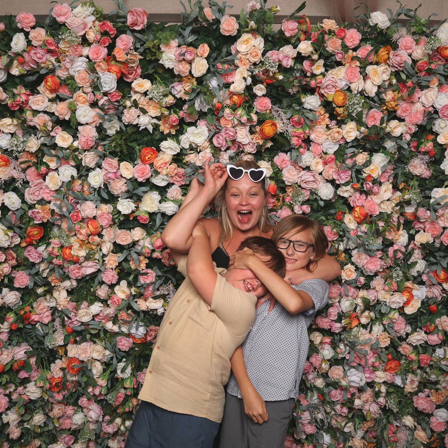 Two people laugh and pose in front of a colorful floral wall backdrop at a celebration.