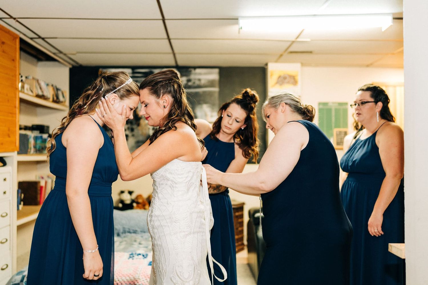 Bridesmaids in navy blue dresses help a bride in a white gown get ready in a cozy room before her wedding.