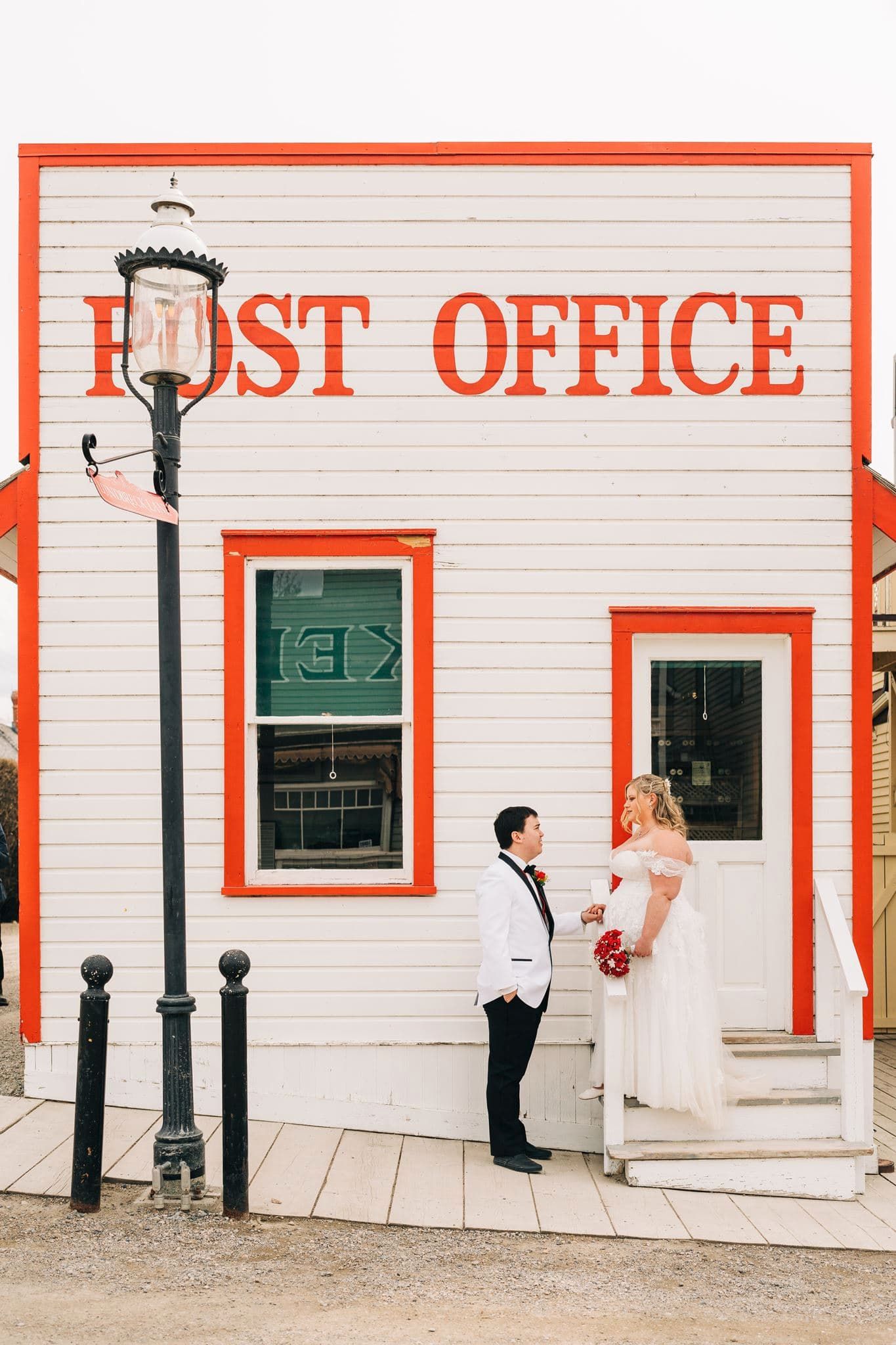 Bride and groom posing outside a charming white and red Post Office building, bride holding a red bouquet.
