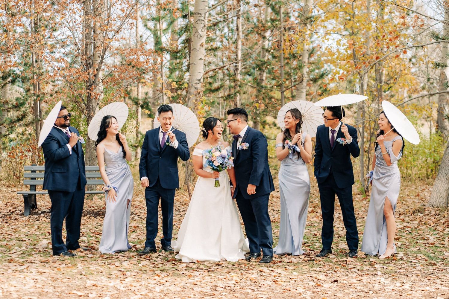 Wedding party in navy suits and silver gowns holding white parasols among autumn foliage, bride and groom sharing a kiss.
