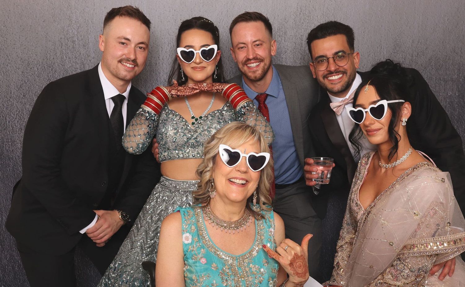 Six people posing in a photo booth, with two women wearing heart-shaped white sunglasses at a festive celebration.