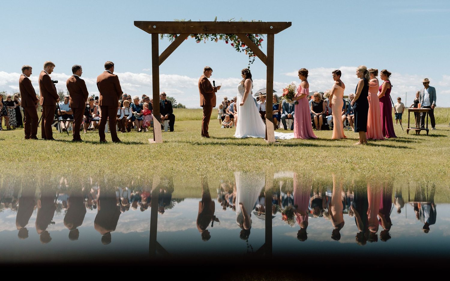 Outdoor wedding ceremony on green lawn with bridal party reflected in still water under rustic wooden arch.