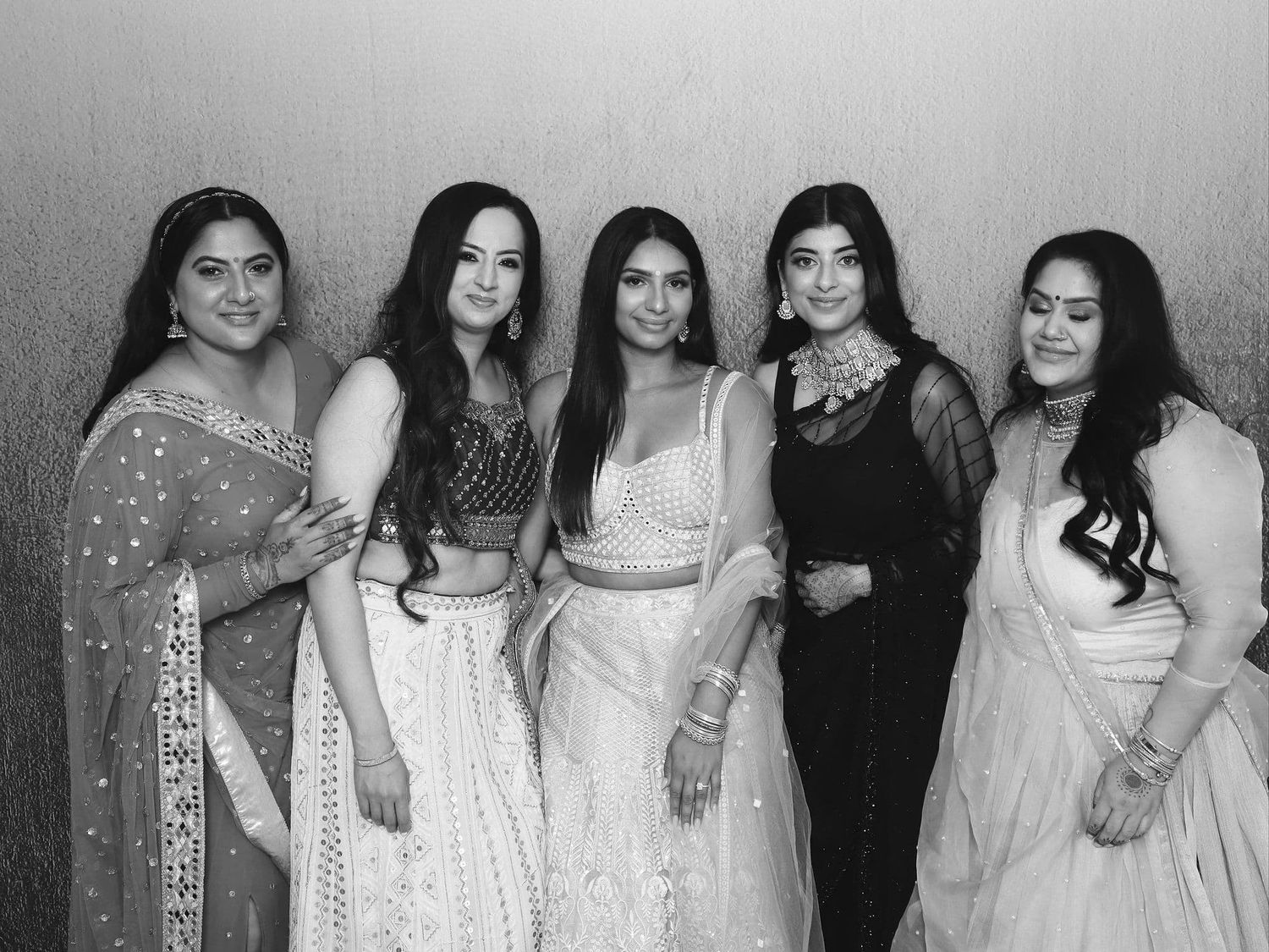 Five women in elegant Indian attire pose together for a black and white photo booth picture.