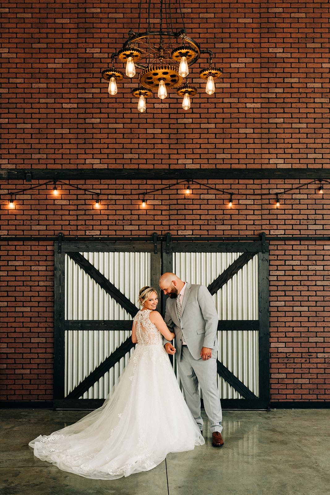 Bride and groom pose romantically under industrial chandelier against brick wall with barn doors at wedding venue.