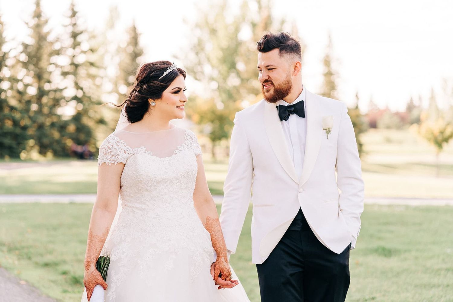 A bride and groom walk hand in hand outdoors, smiling at each other in elegant white wedding attire on a sunny day.