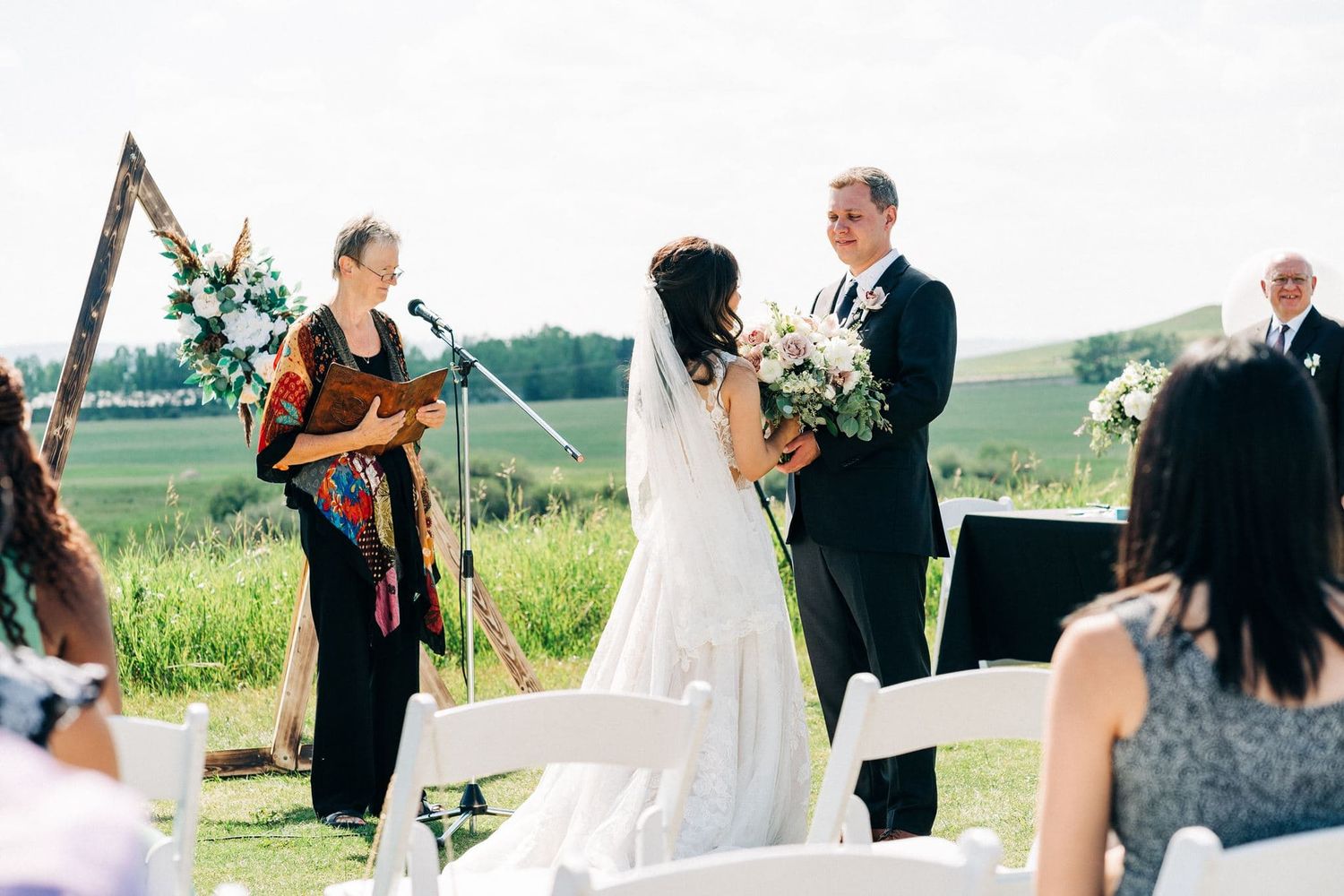 Outdoor wedding ceremony with bride and groom exchanging vows before officiant, set against lush green countryside backdrop.