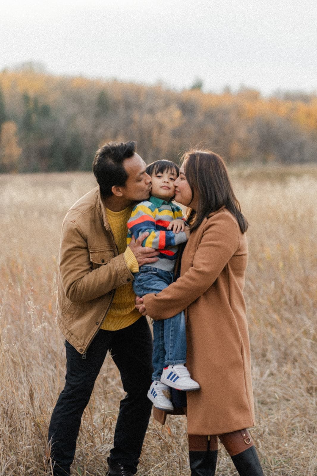 A couple kisses their toddler on the cheeks in an autumn field, the child wearing a colorful striped sweater.