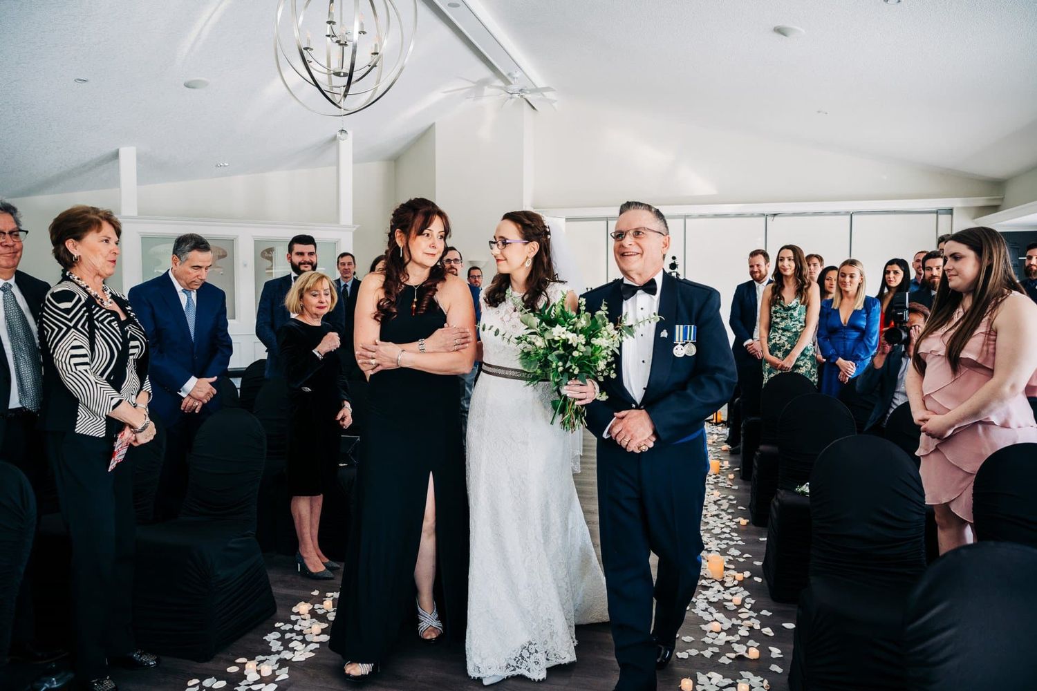 Bride walks down the aisle with family at an elegant indoor wedding ceremony.