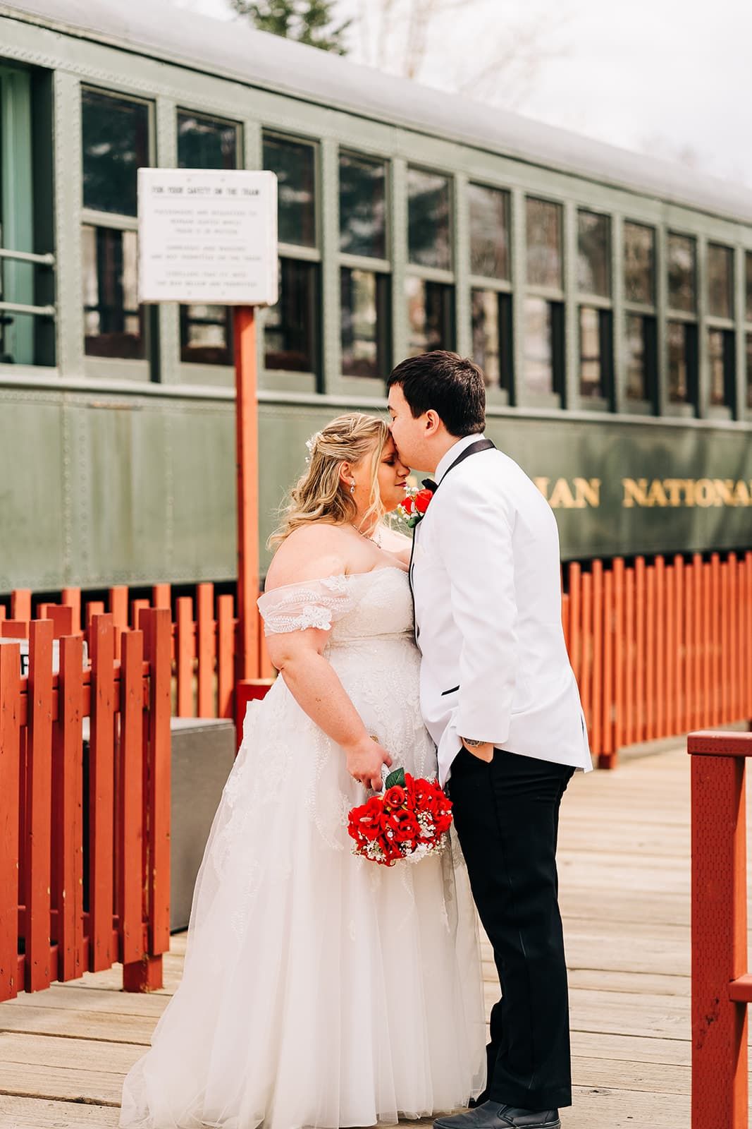 A bride and groom share a kiss on a wooden platform beside a vintage green train car, bride holding red bouquet.
