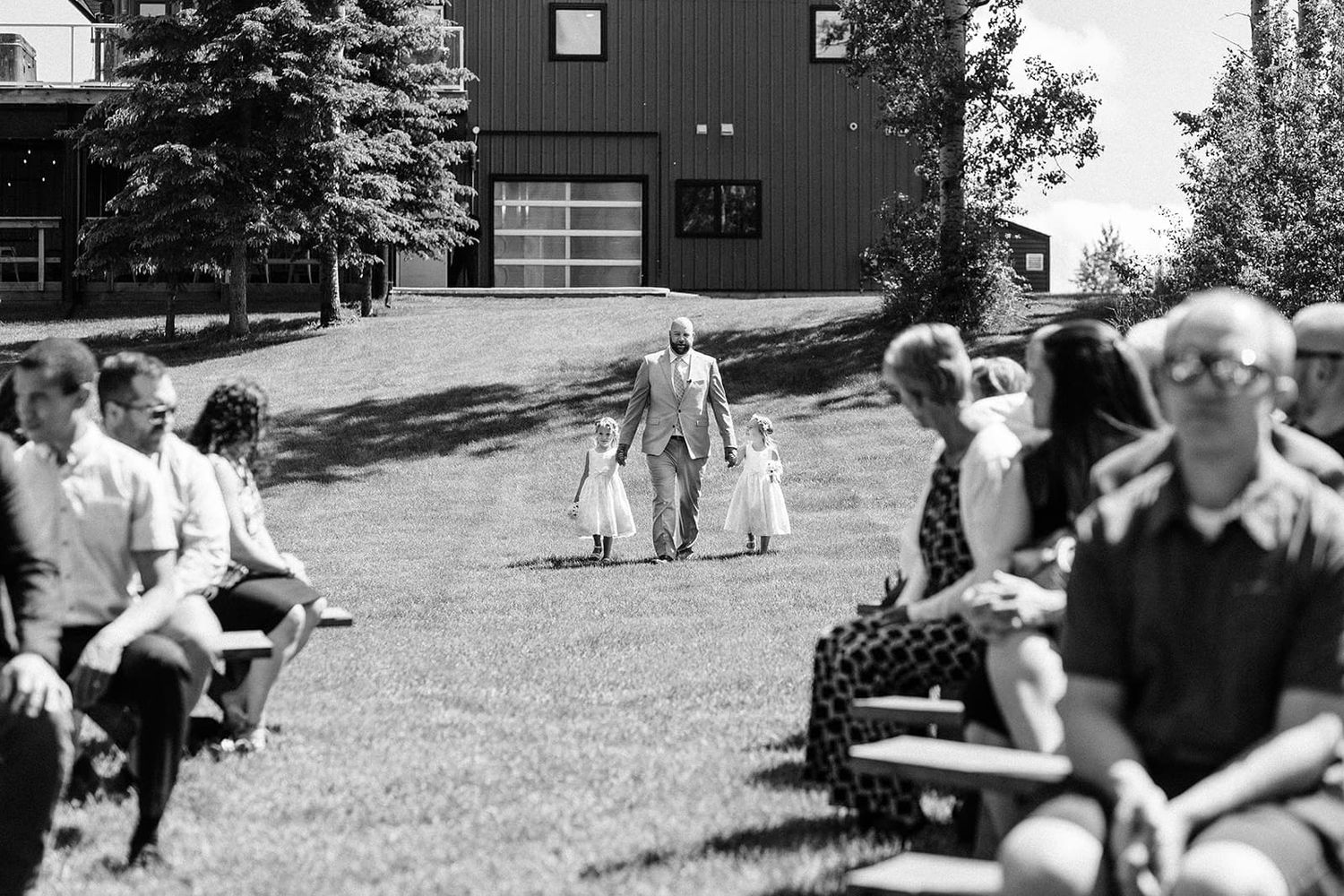 Black and white outdoor wedding ceremony with flower girls walking down grassy aisle flanked by seated guests.
