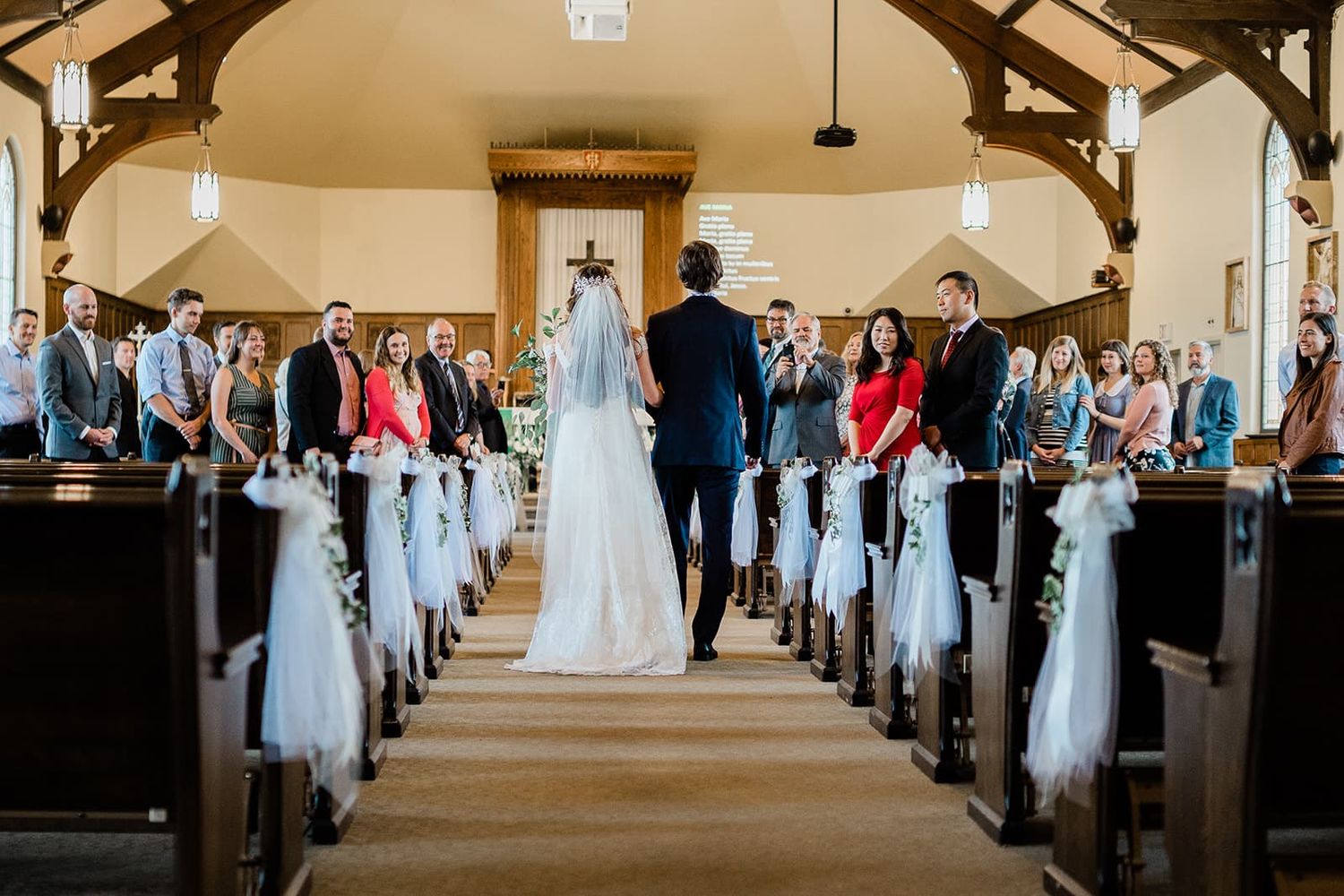 Bride walking down the aisle in a white gown and veil in a wooden church filled with wedding guests.